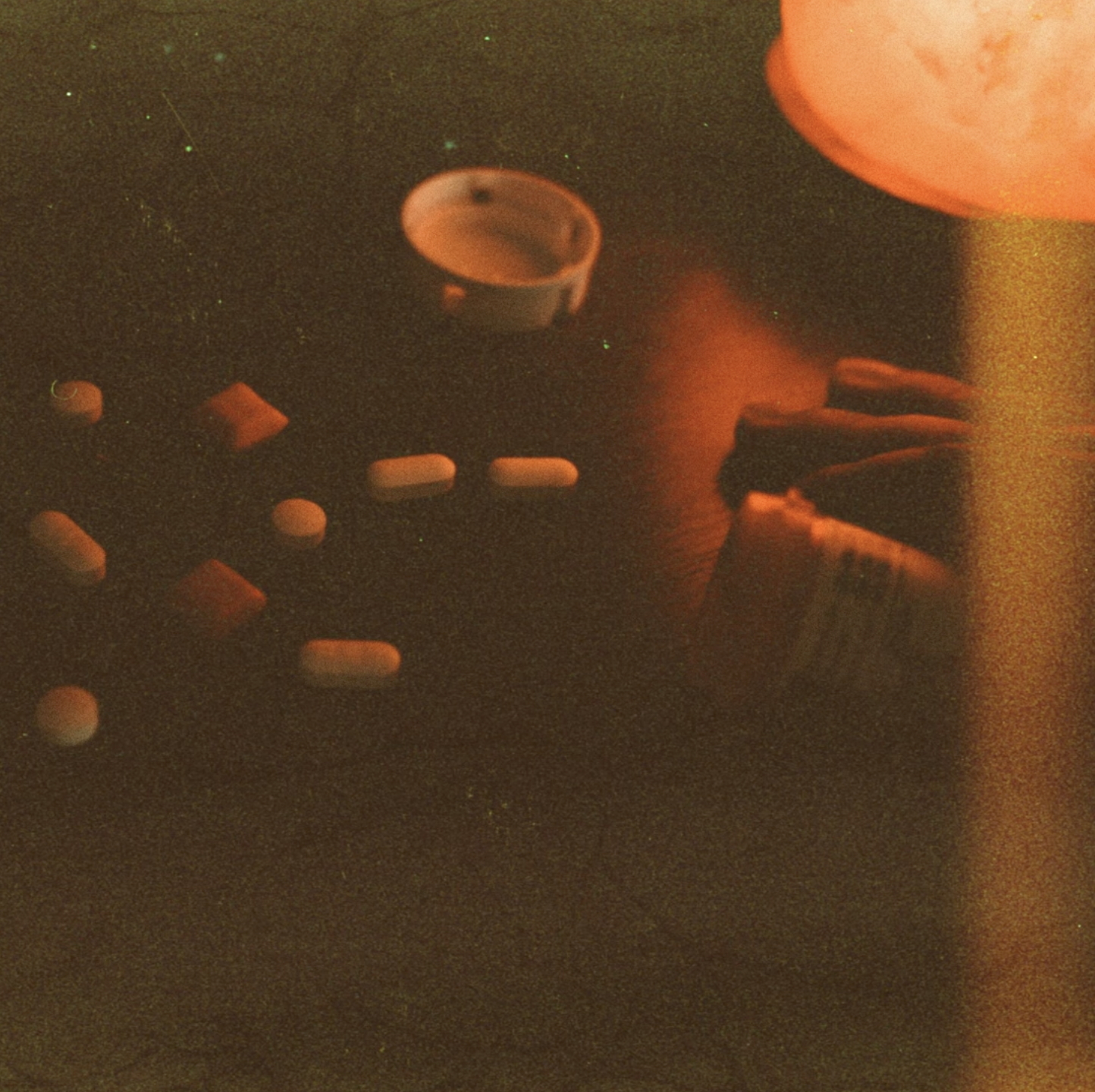 Close-up of a dark surface with scattered pills and a glass of water, illuminated by an orange light.