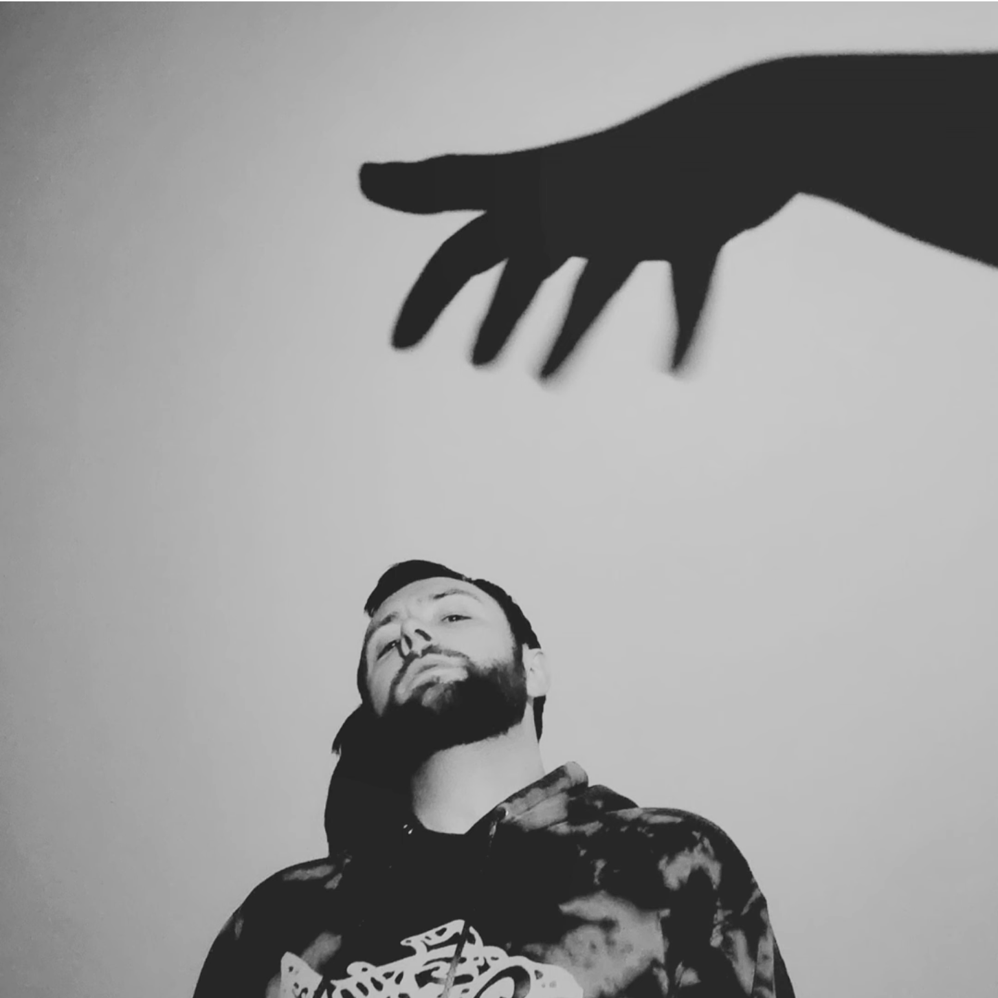 Black and white photograph of a man with a beard looking up, with a large shadow of a hand and fingers cast above him on the wall.