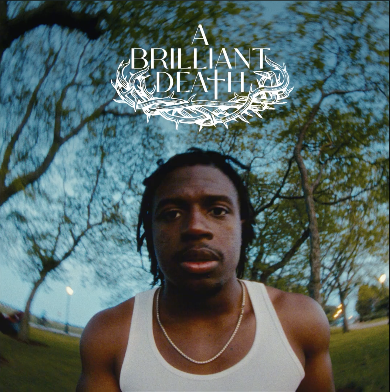 A young man with dreadlocks wearing a white tank top and a silver chain, standing outdoors in a park with trees and a blue sky in the background, with the title 'A Brillant Death' overlayed at the top.