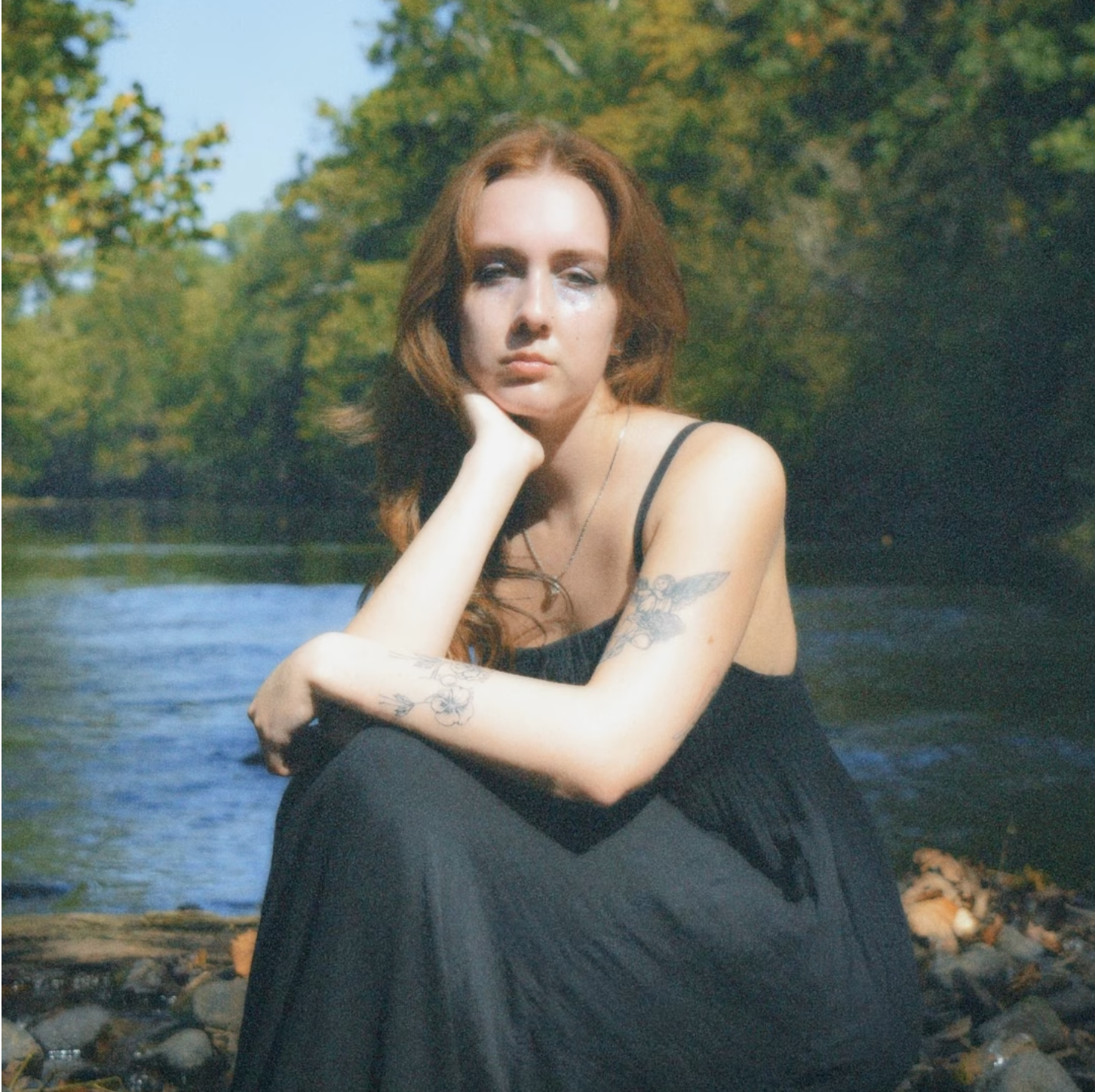 A young woman with red hair and tattoos, sitting outdoors by a river with green trees in the background.