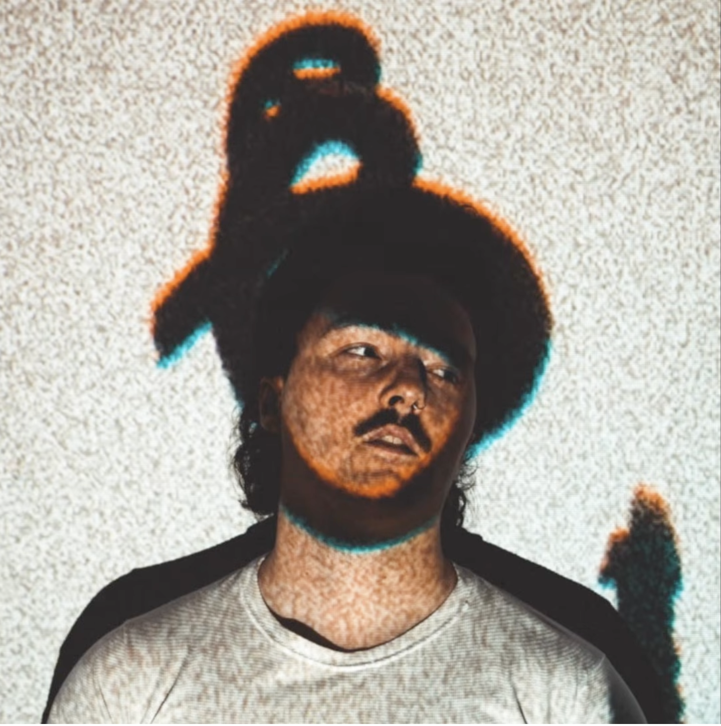 A man with dark wavy hair, a mustache, and a beard wearing a gray t-shirt with black sleeves, standing against a textured white wall, with a fuzzy image effect.