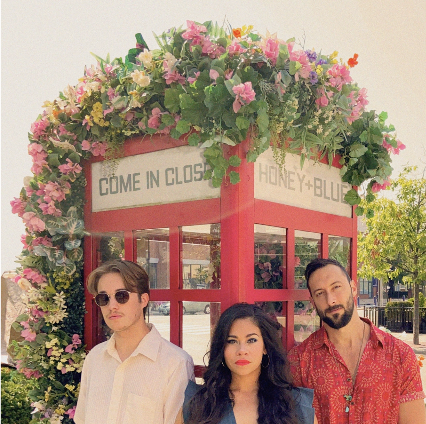 Group of three adults standing in front of a red phone booth decorated with pink, yellow, and white flowers, with a sign that reads 'Come in close, honey + blue'. It is a sunny day with trees and a street visible in the background.