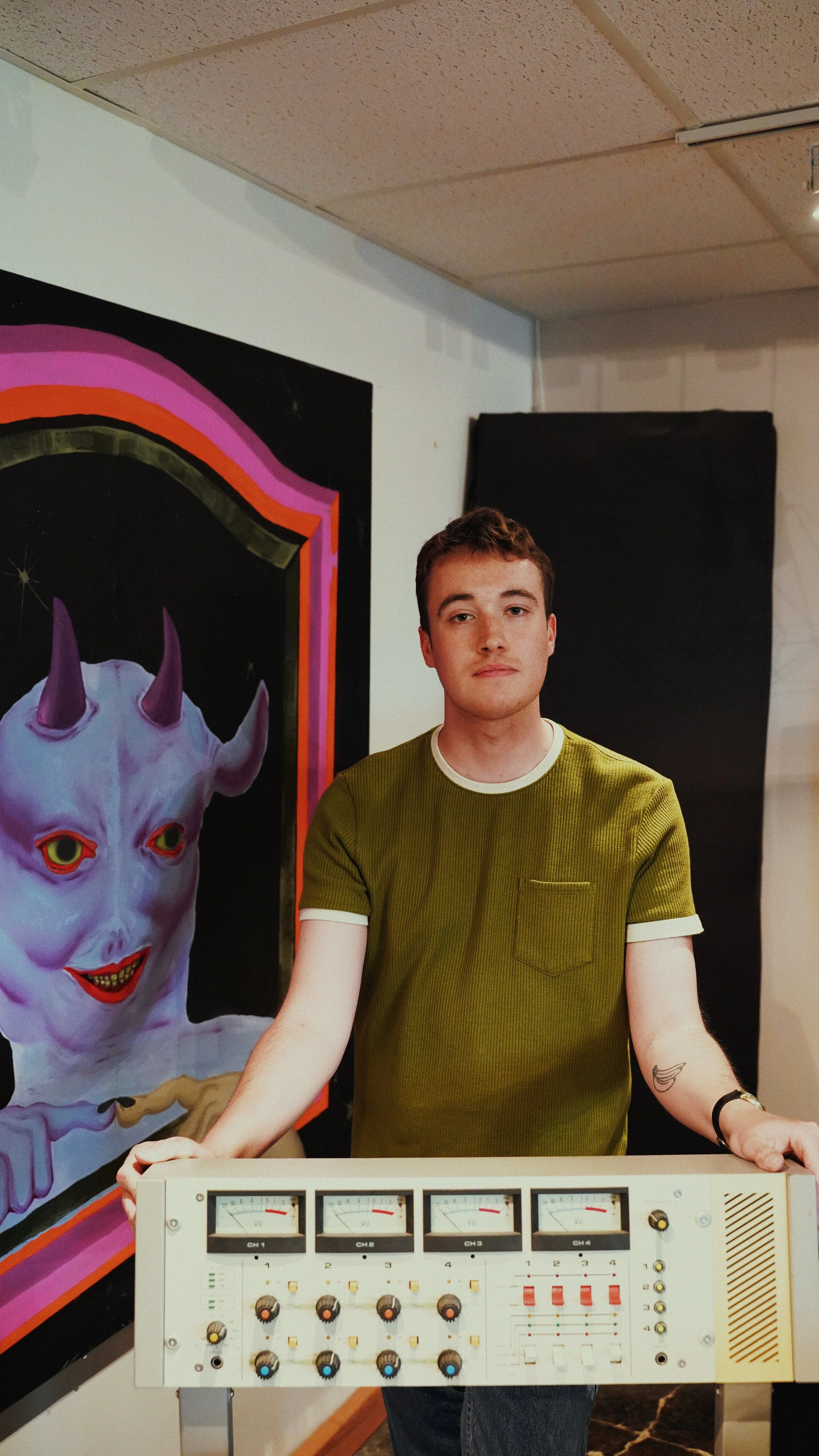 A young man standing behind a vintage reel-to-reel machine with four meters, knobs, and switches, wearing a green T-shirt with white cuffs, in a room with a painting of a purple cartoonish devil with horns and a smiling face in the background.