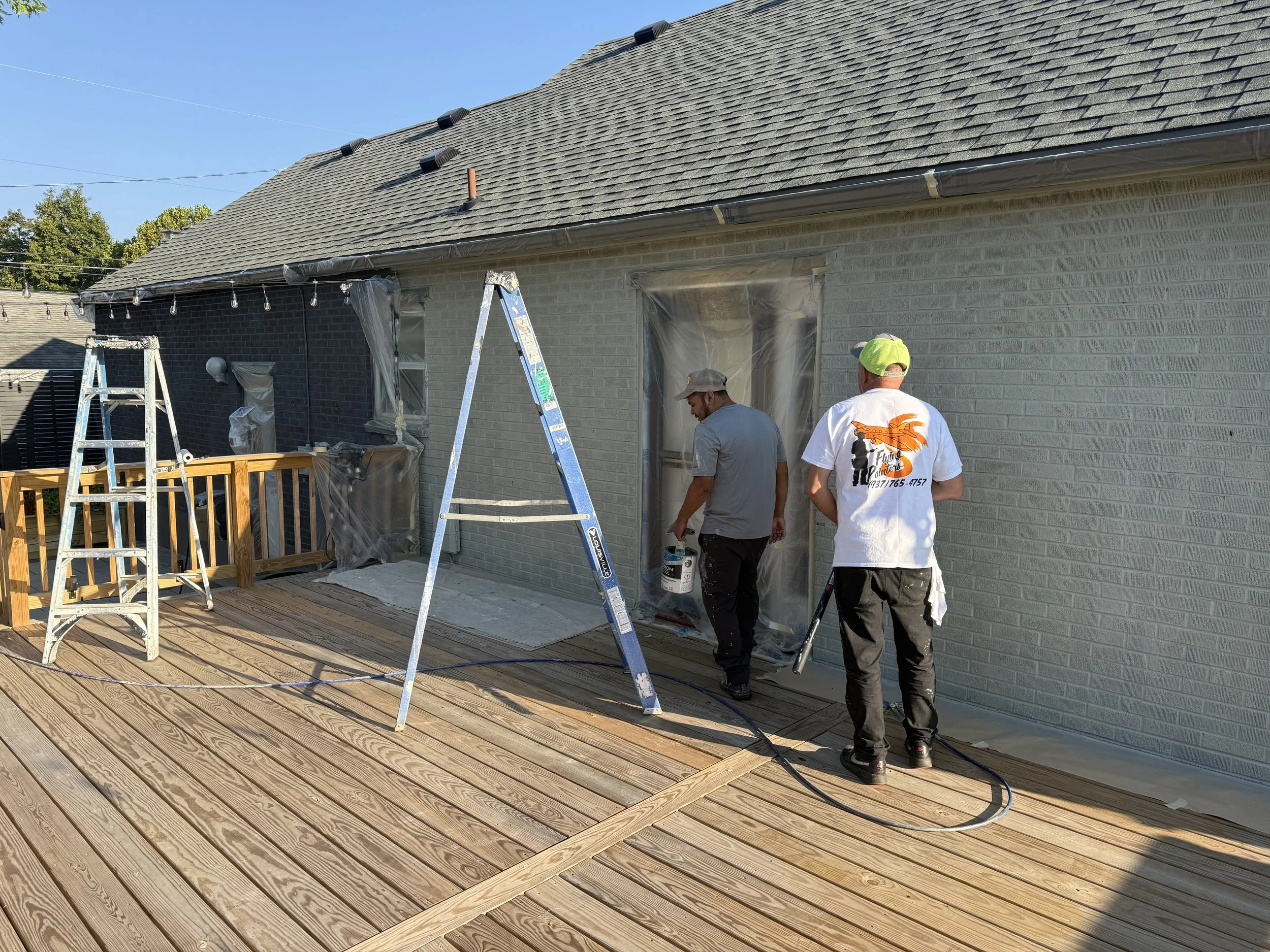 Two men are working on an exterior house renovation with scaffolding and ladders. The house has a new shingled roof and a light-colored brick exterior. Plastic coverings protect the windows, and a string of lights hangs under the roof. One man wears a white T-shirt and a green cap, the other a gray shirt and a beige cap.
