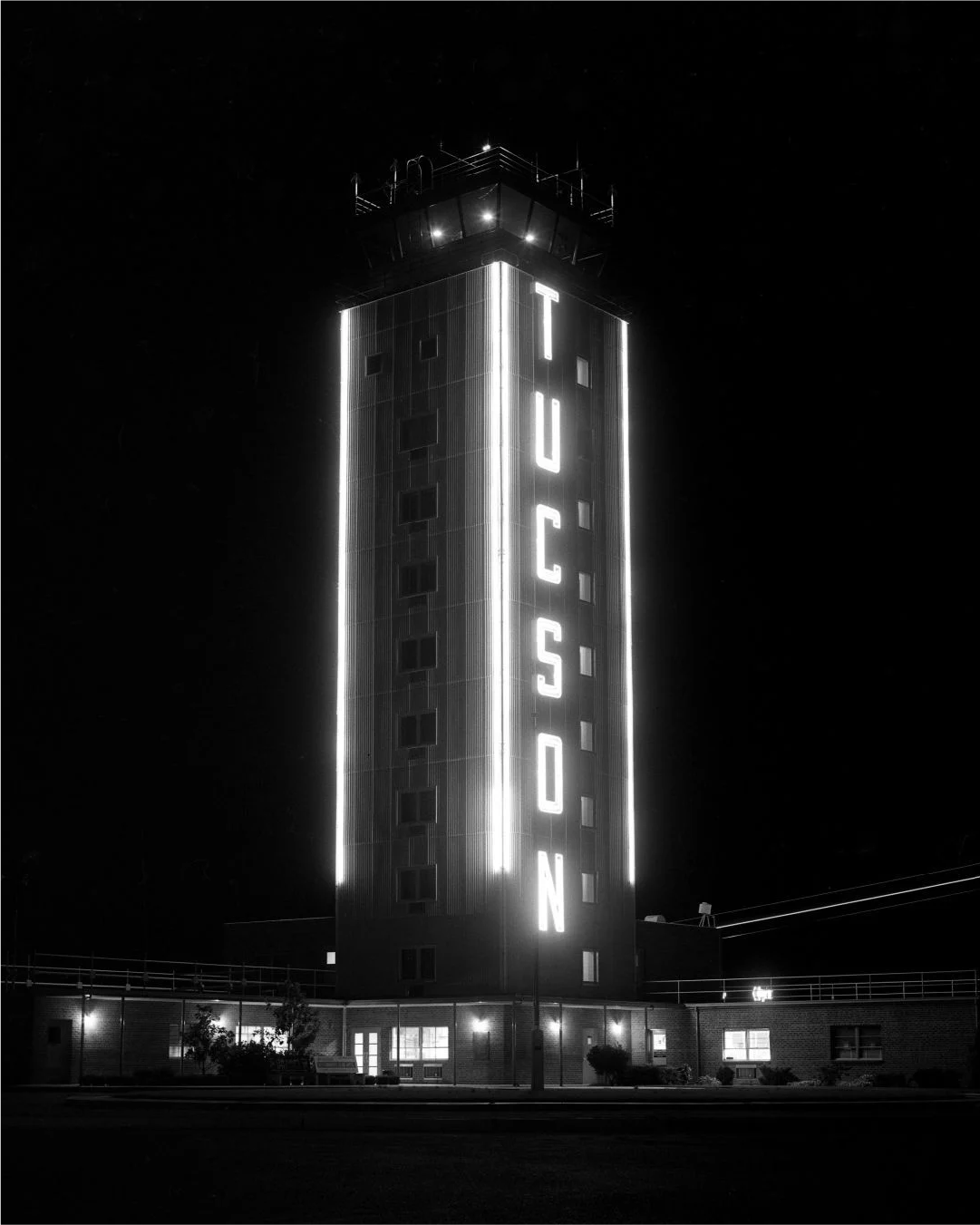 Nighttime black and white photo of a tall building with bright vertical lights and a large illuminated sign that reads 'TUCSON' on the side.