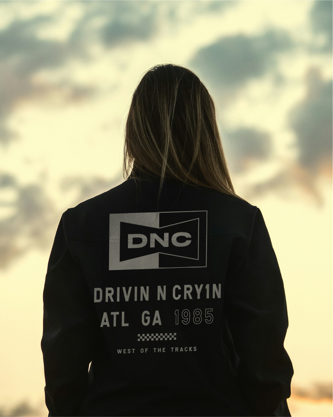 Back of a person with long hair wearing a black jacket with DNC logo and text, standing outdoors at sunset with cloudy sky in background.