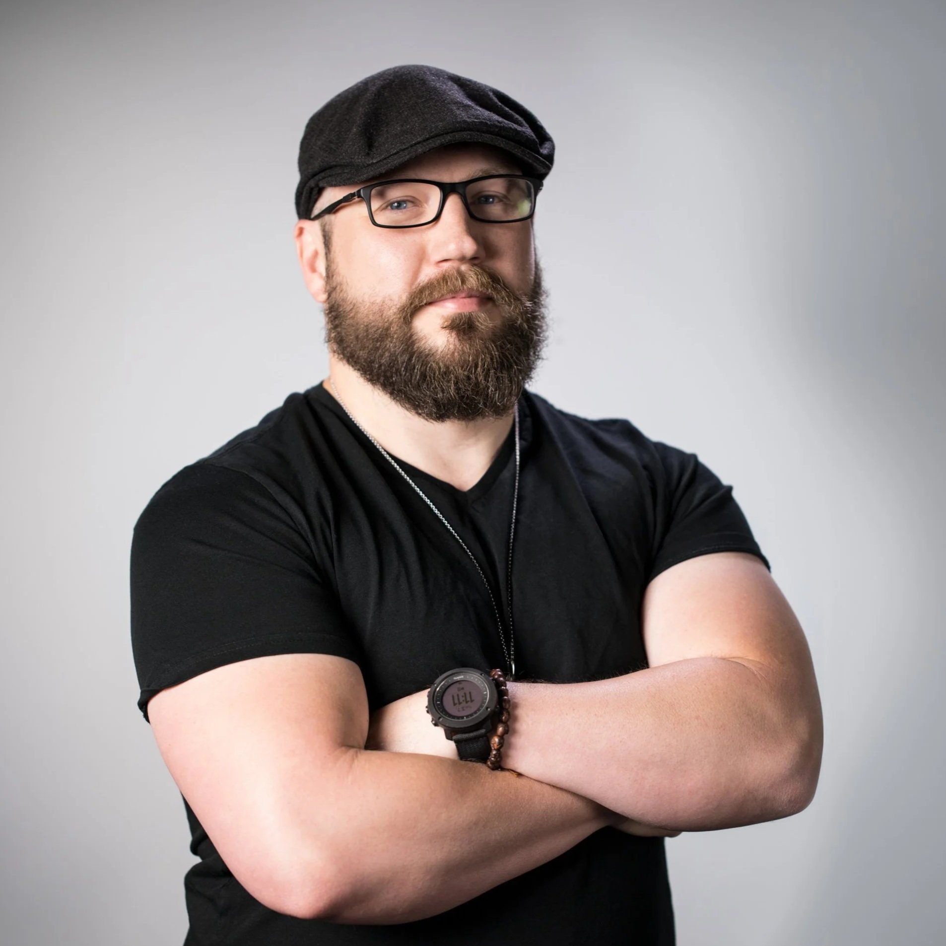 Aaron James Gresham: A bearded man wearing glasses, a black cap, a black shirt, and a watch, standing with arms crossed against a plain light gray background.