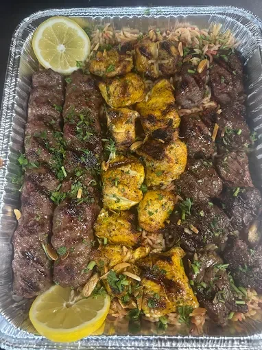 Baked skewers of grilled beef and chicken served with rice, herbs, and lemon wedges in a foil container.