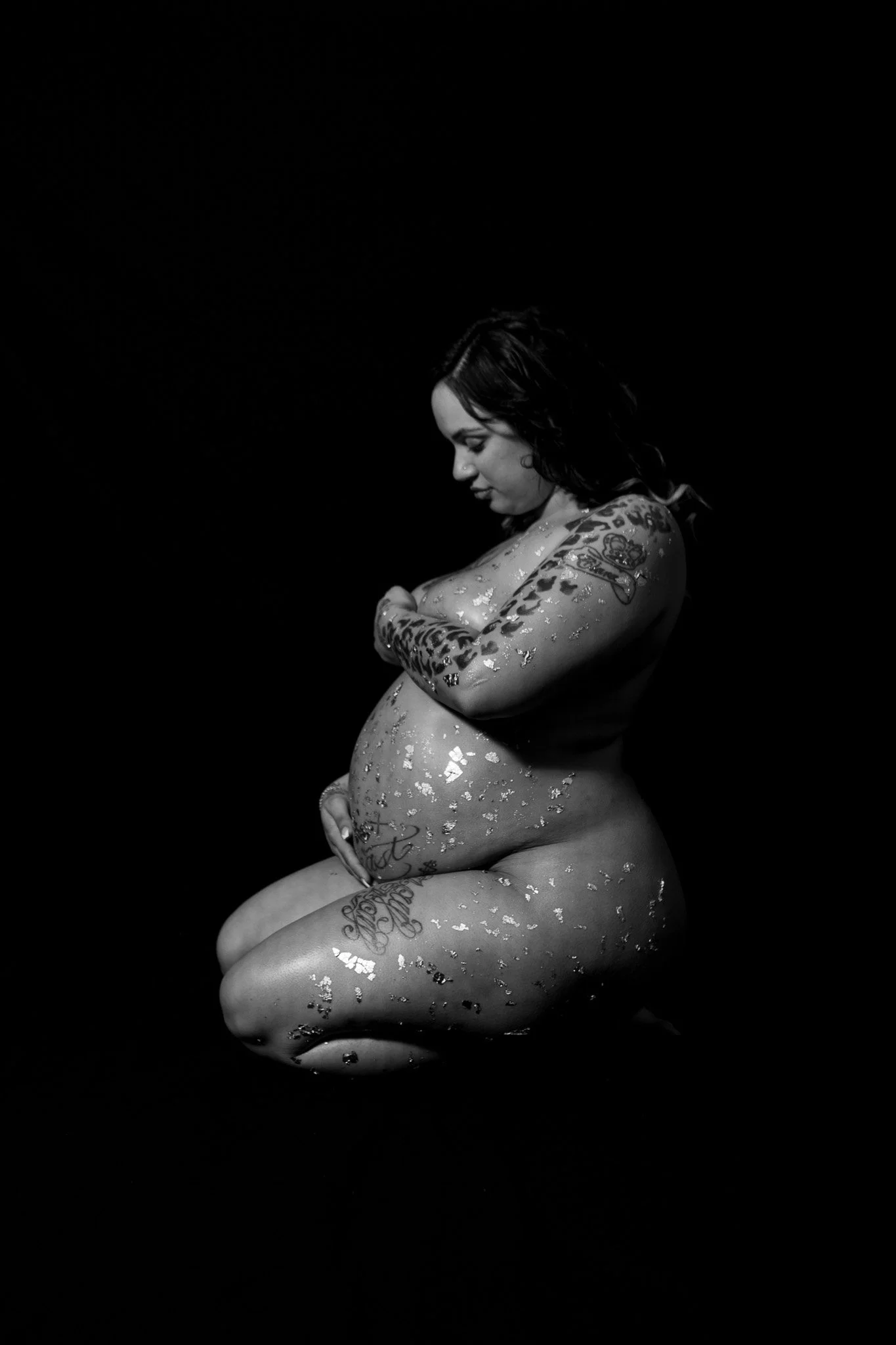 Black and white photo of a pregnant woman with tattoos, sitting with her eyes closed, gently resting her hands on her belly, against a dark background.