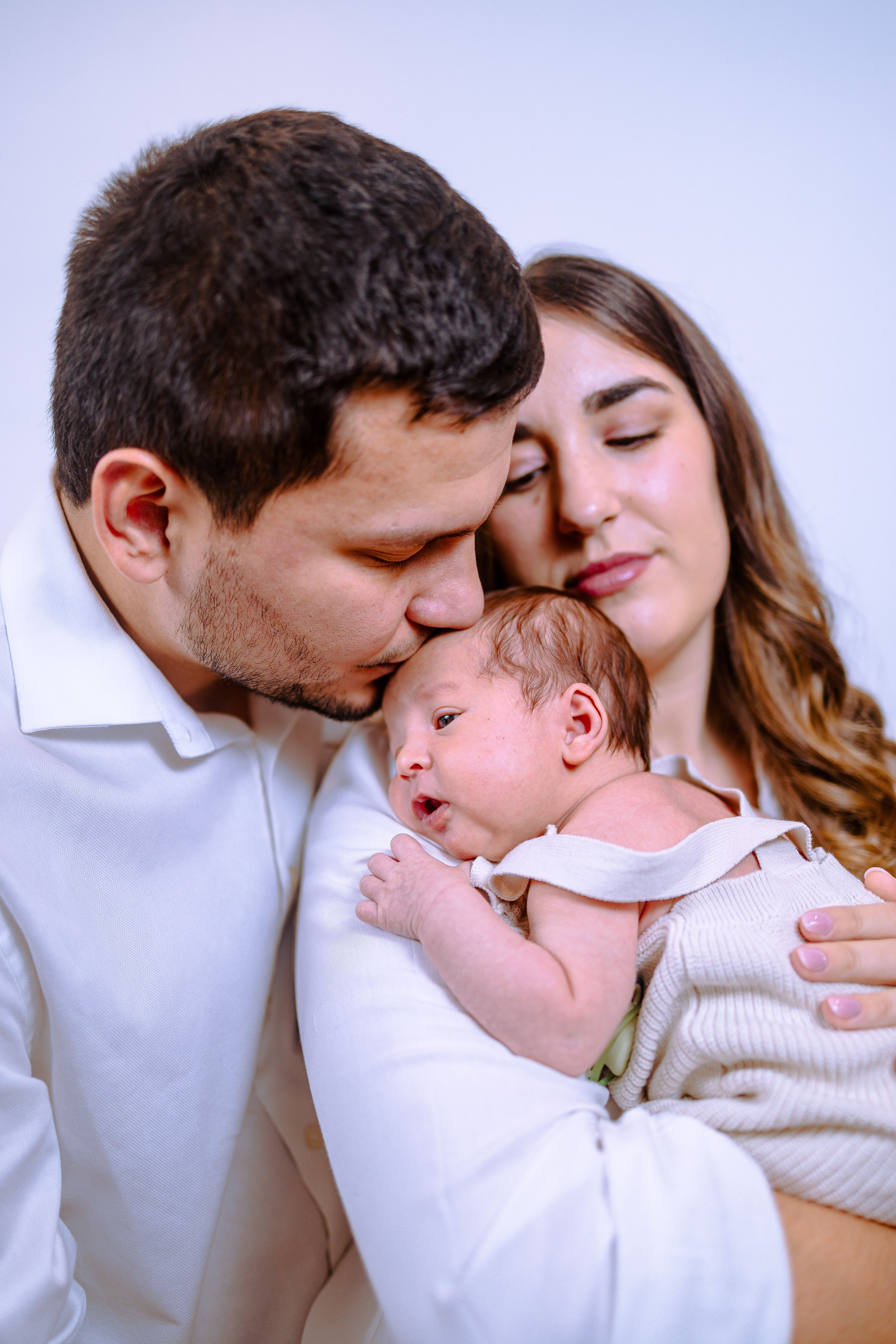 A family of three, a man, woman, and baby, sharing a tender moment. The man is kissing the baby's forehead, the woman is gently holding the baby, all with closed eyes and peaceful expressions.