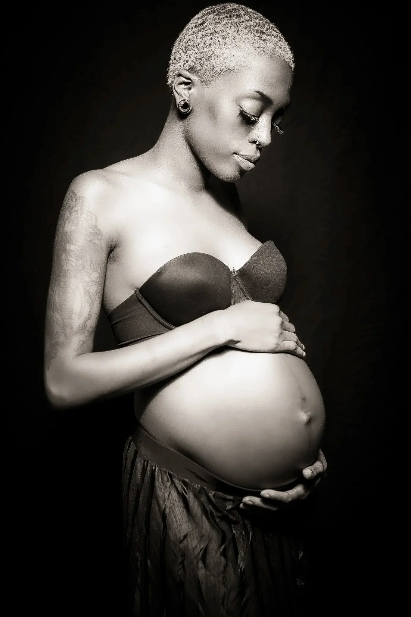 A pregnant woman with short blonde hair, tattoos on her arms, and earrings, wearing a strapless top and patterned pants, cradling her belly with her hands and looking down, in black and white against a dark background.