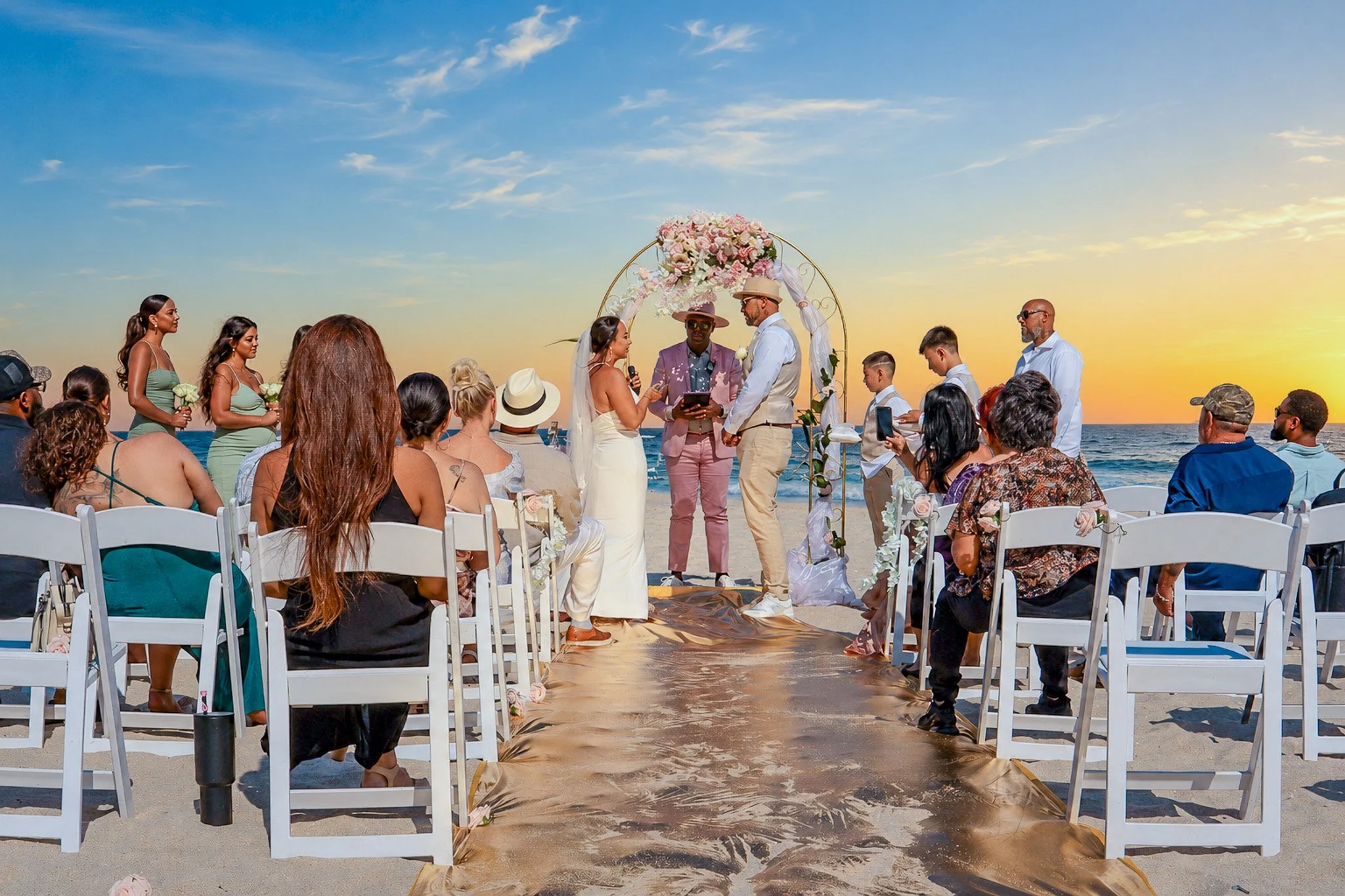 Beachside wedding ceremony with an arch decorated with pink flowers, officiant, couple exchanging vows, guests seated on white chairs, sunset over the ocean in the background.