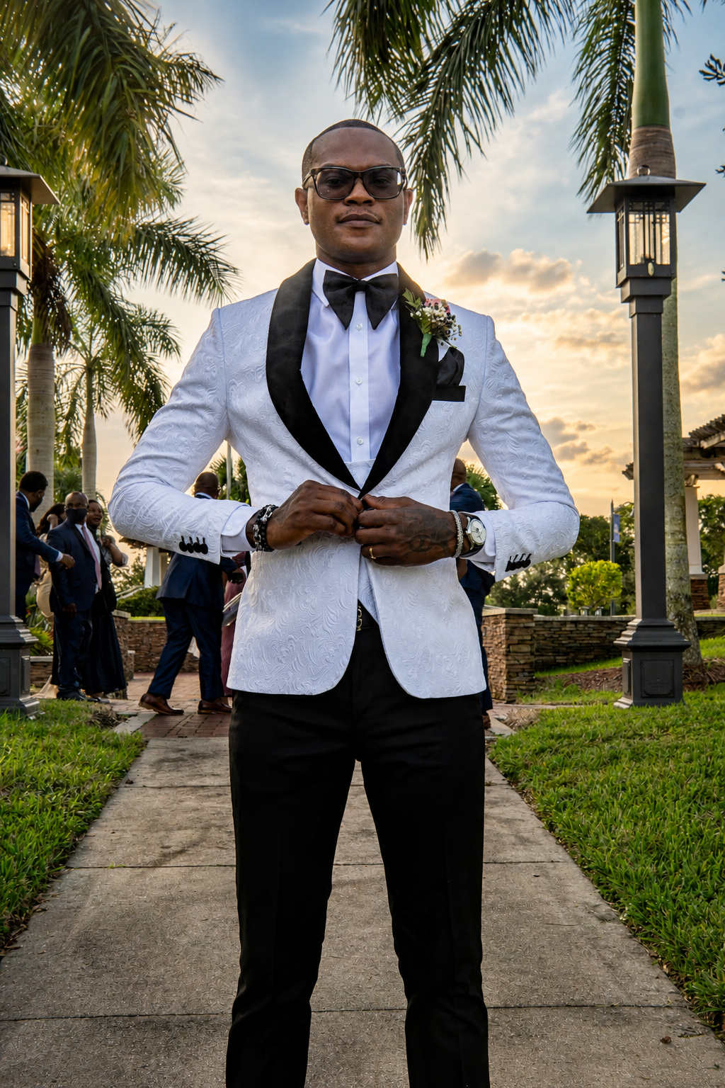 A man in a white tuxedo with a black lapel and black bow tie adjusting his jacket, standing outdoors during sunset with palm trees and other people in the background.