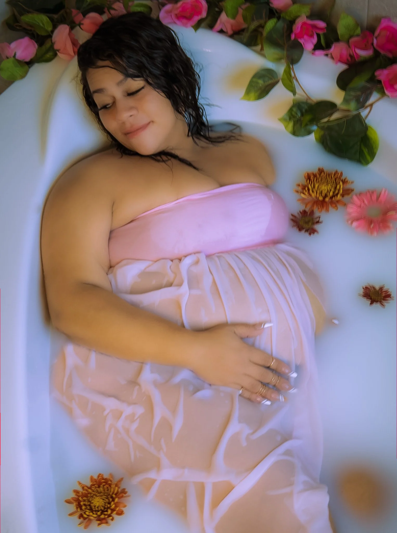 A woman with wet, dark hair relaxing in a white bathtub, surrounded by pink and orange flowers, with her eyes closed and a serene expression.