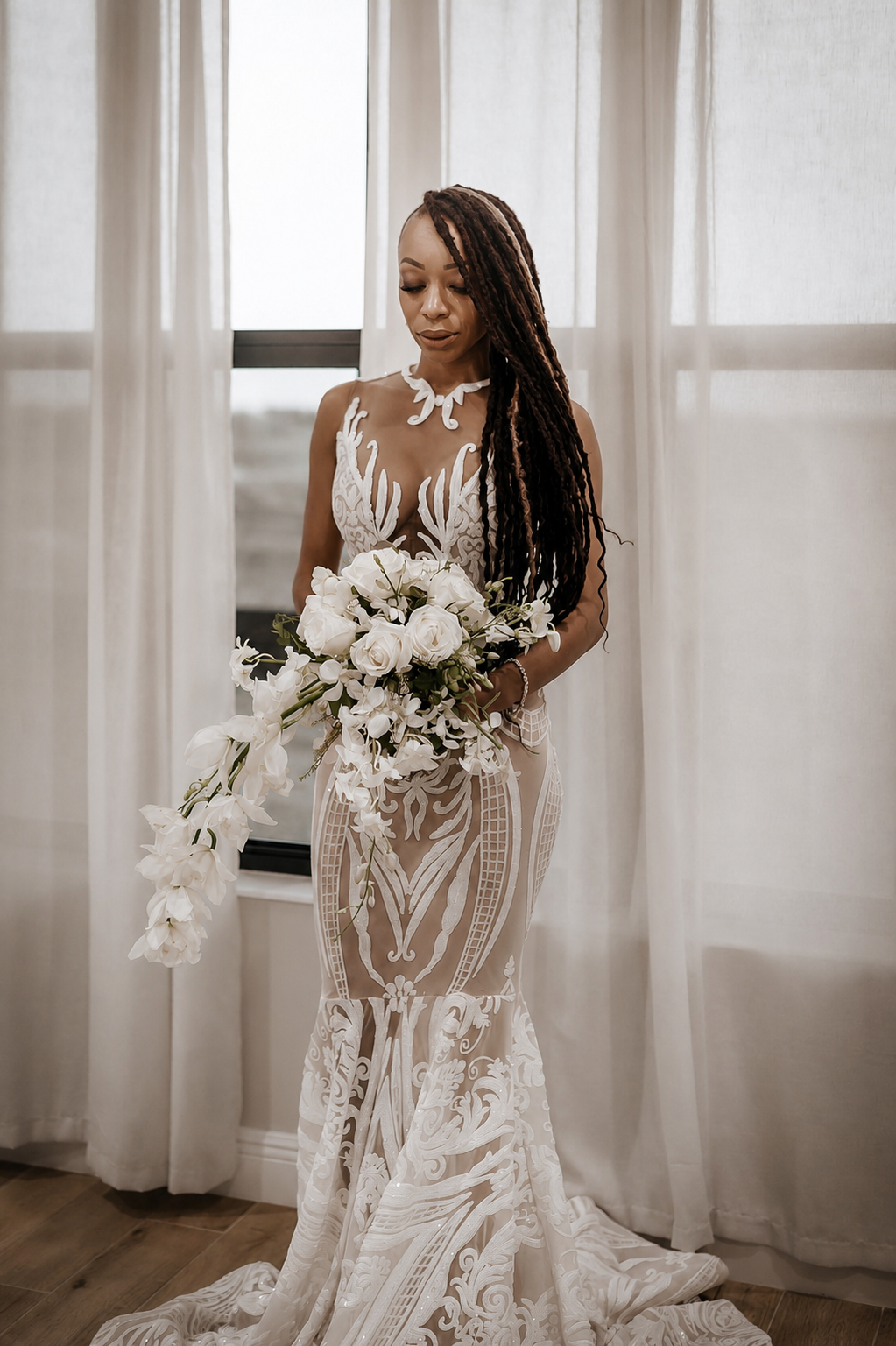 A woman in an elegant white lace wedding dress holding a cascading bouquet of white roses and orchids inside a room with sheer curtains.