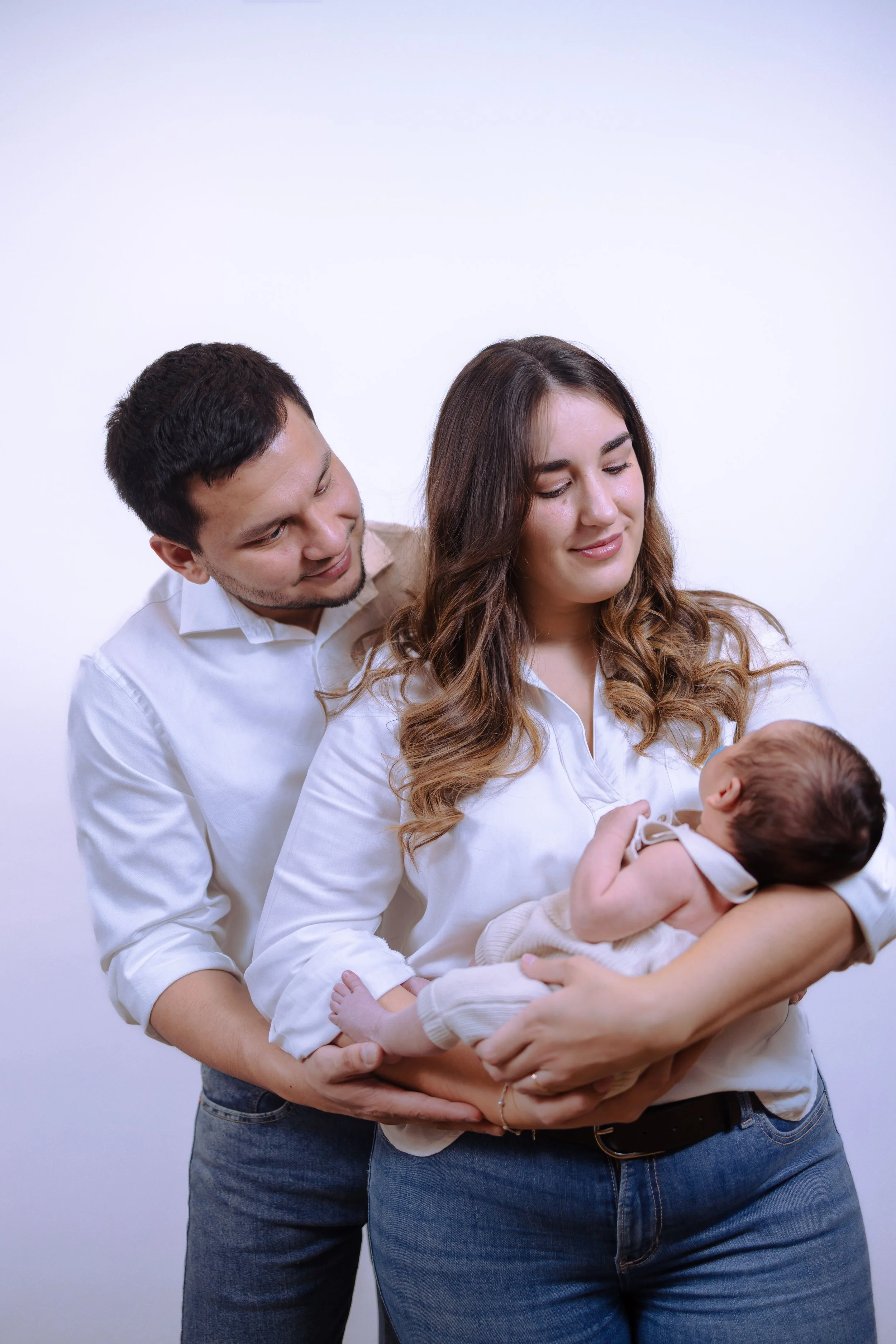 A family of three, a man, woman, and baby, standing against a plain white background. The man and woman are dressed in white shirts and are holding the baby, who is wearing beige pants and a white top, in their arms.