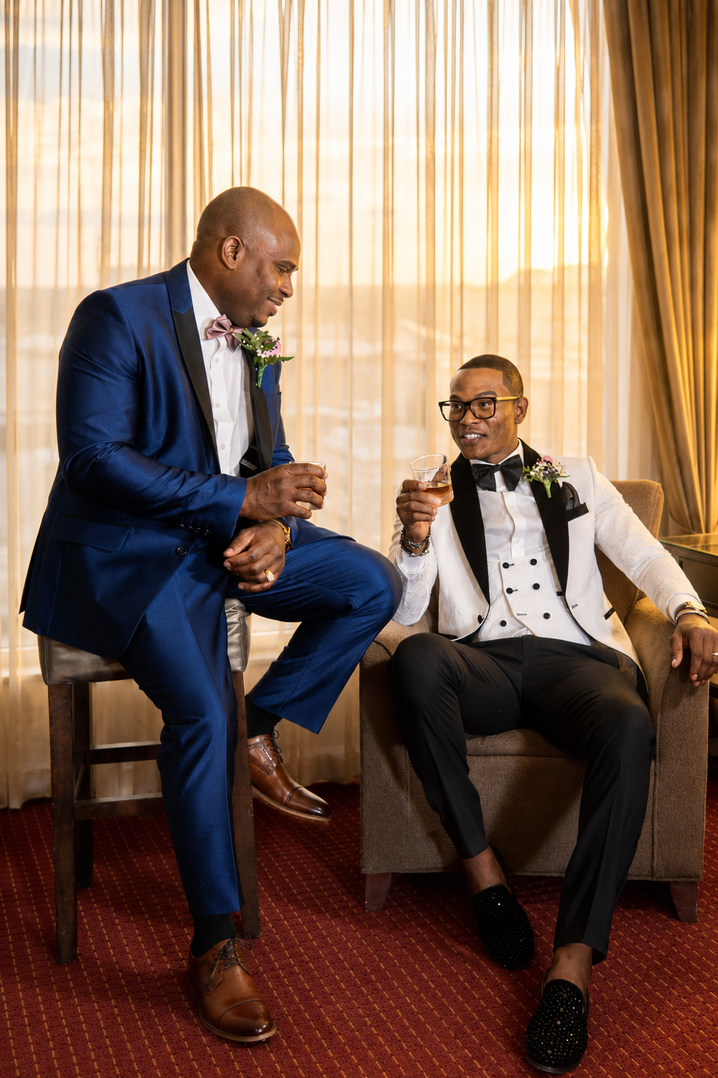 Two men in tuxedos enjoying drinks and conversation in a warmly lit room with curtains.