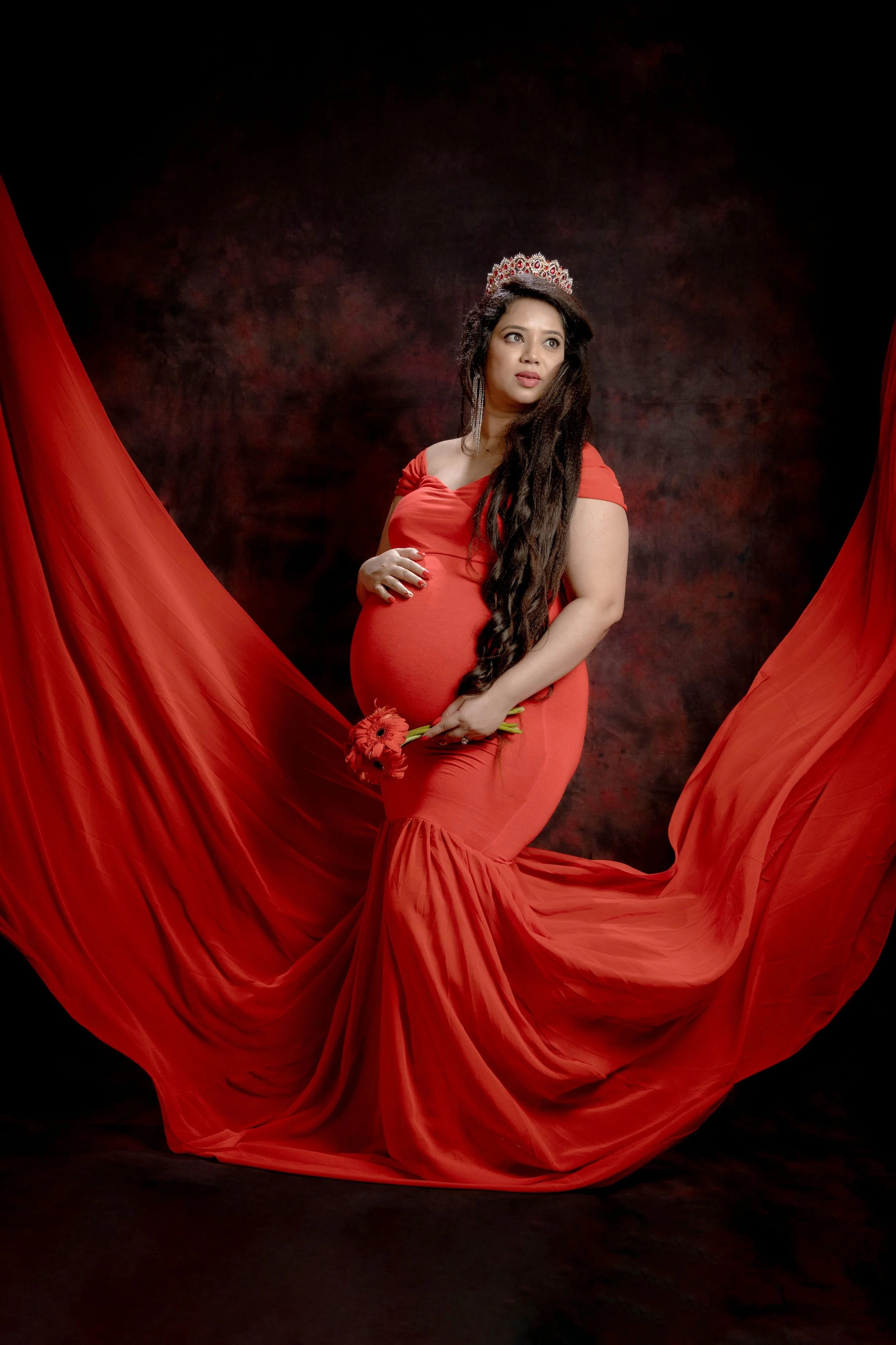 A pregnant woman in a red dress with flowing fabric, wearing a silver tiara and earrings, holding red flowers, posed against a dark, textured background.