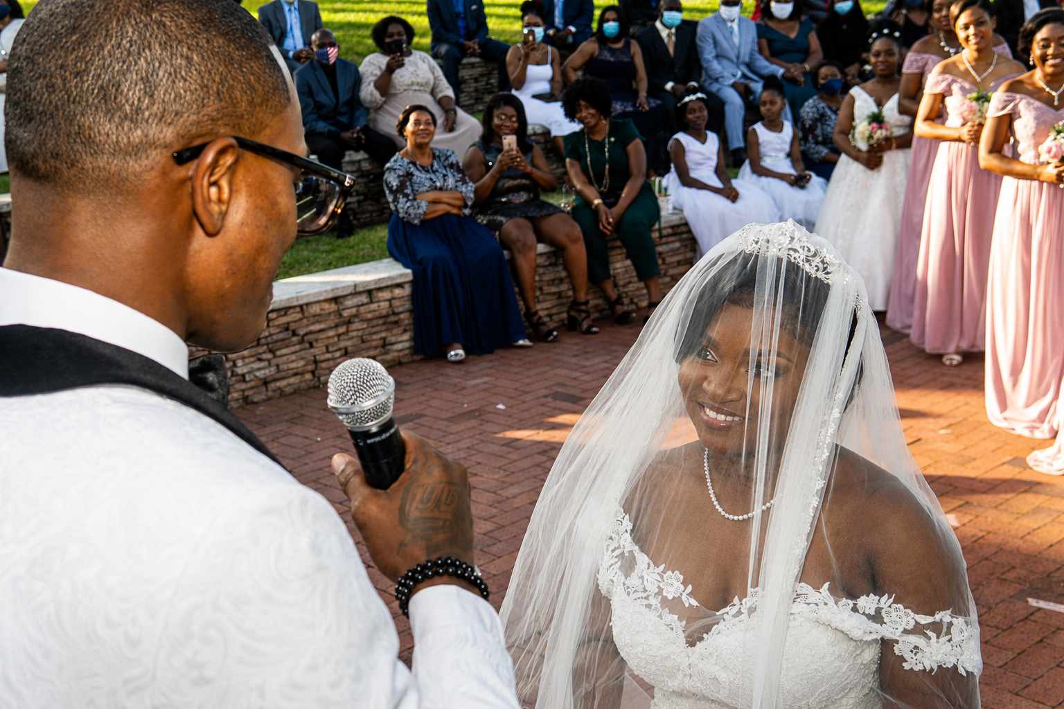 A groom holding a microphone faces a smiling bride wearing a veil at their wedding ceremony outdoors, with wedding guests seated and standing in the background.