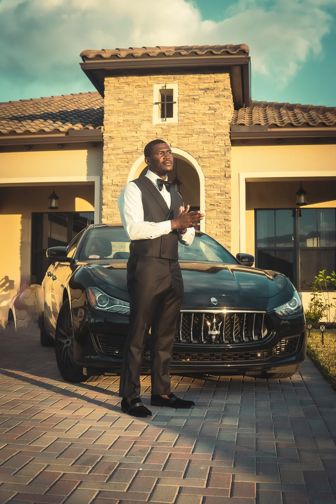 A man dressed as a butler standing in front of a black Maserati sports car outside a modern beige house with stone accents and a tiled roof.