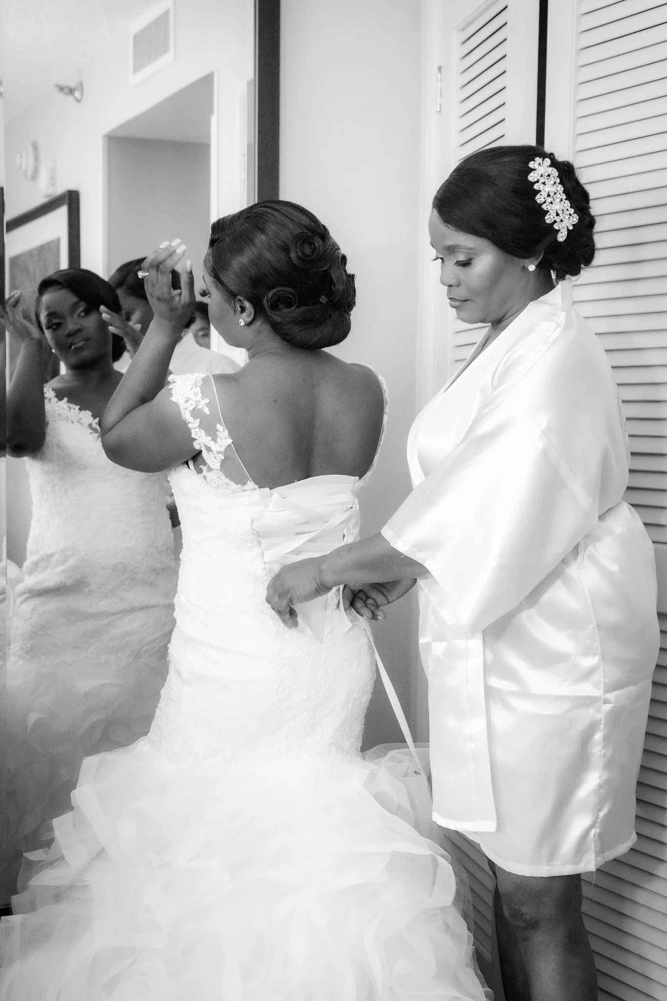 Women in wedding dresses getting ready, with one woman helping another with her dress, in black and white.