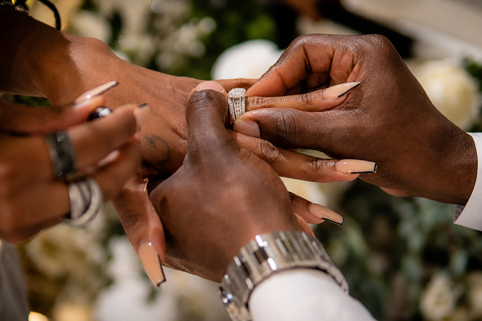 Close-up of a wedding ceremony where a person is placing a diamond ring on another person's finger, with hands adorned with rings and a wristwatch, among blurred flowers in the background.