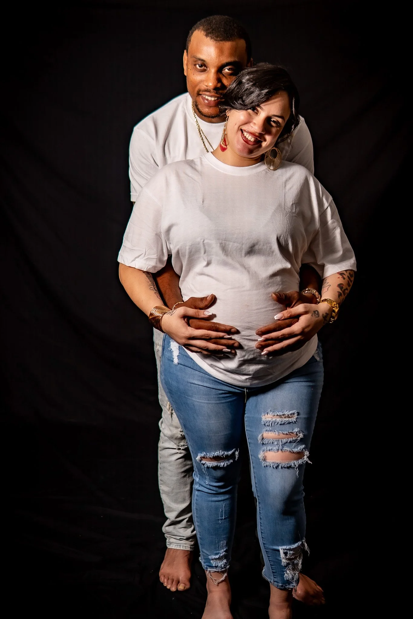 A man and a woman standing together against a black background, with their hands on her pregnant belly. The man is behind the woman, with his arms around her. The woman is smiling and wearing distressed jeans and earrings, while the man is wearing a 
