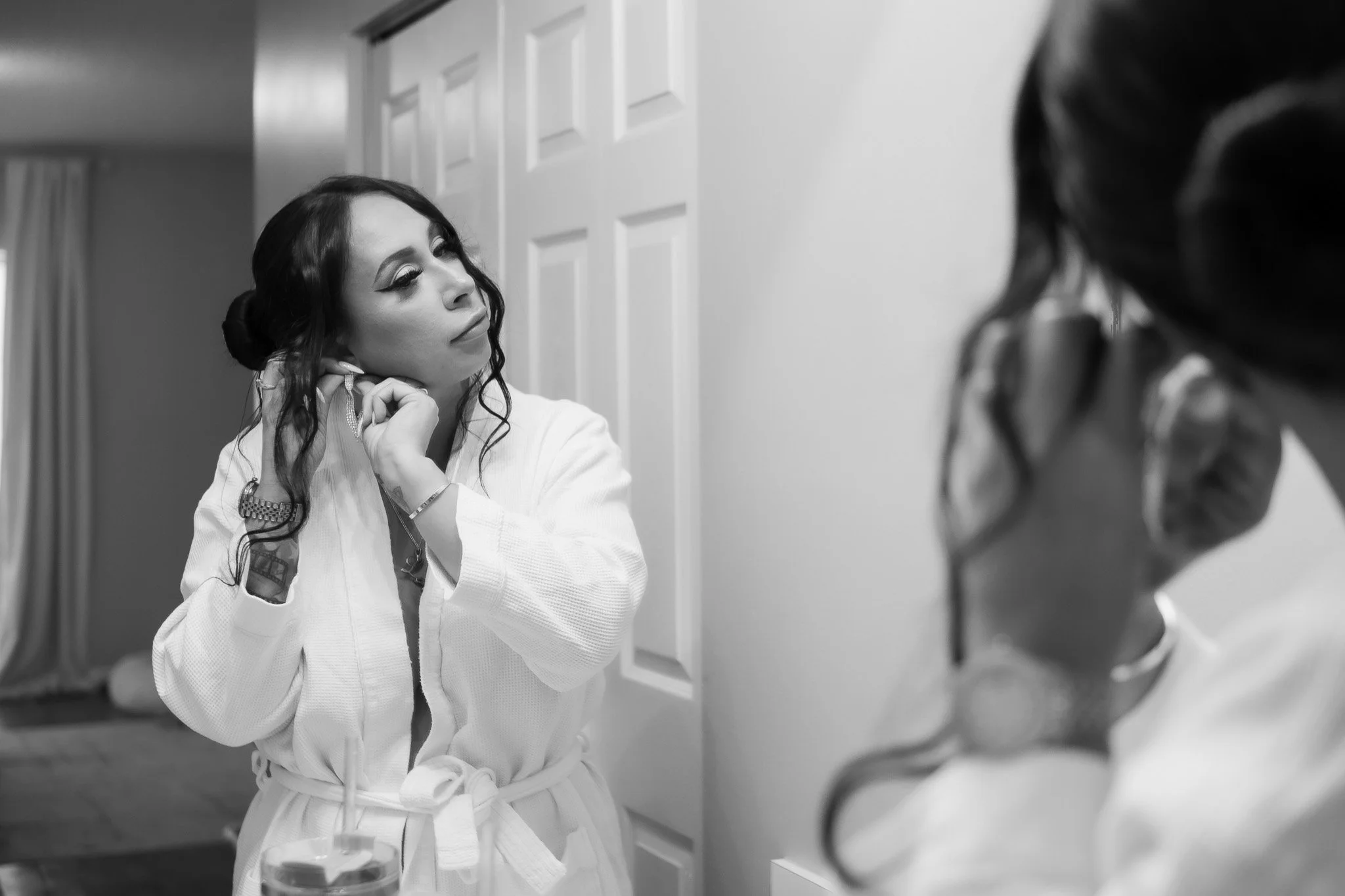 A woman with long dark hair and makeup, wearing a white martial arts gi, is looking at herself in a mirror while adjusting an earring.