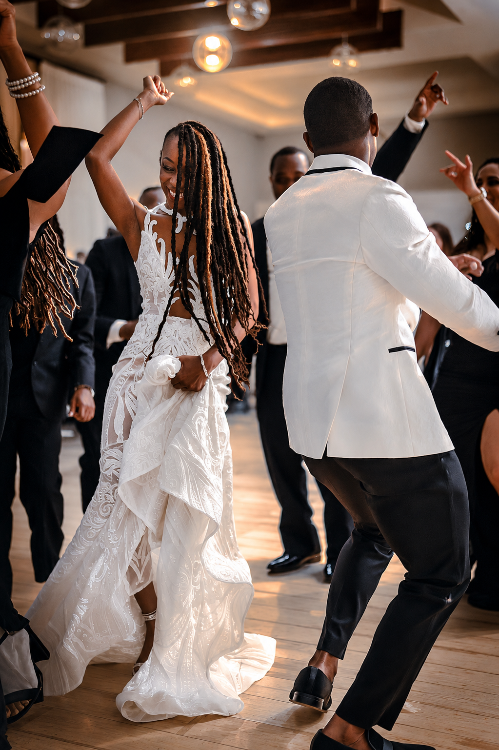 People dancing at a celebration or wedding reception, with a woman in a white lace dress and a man in a light-colored tuxedo dancing among others in formal attire.