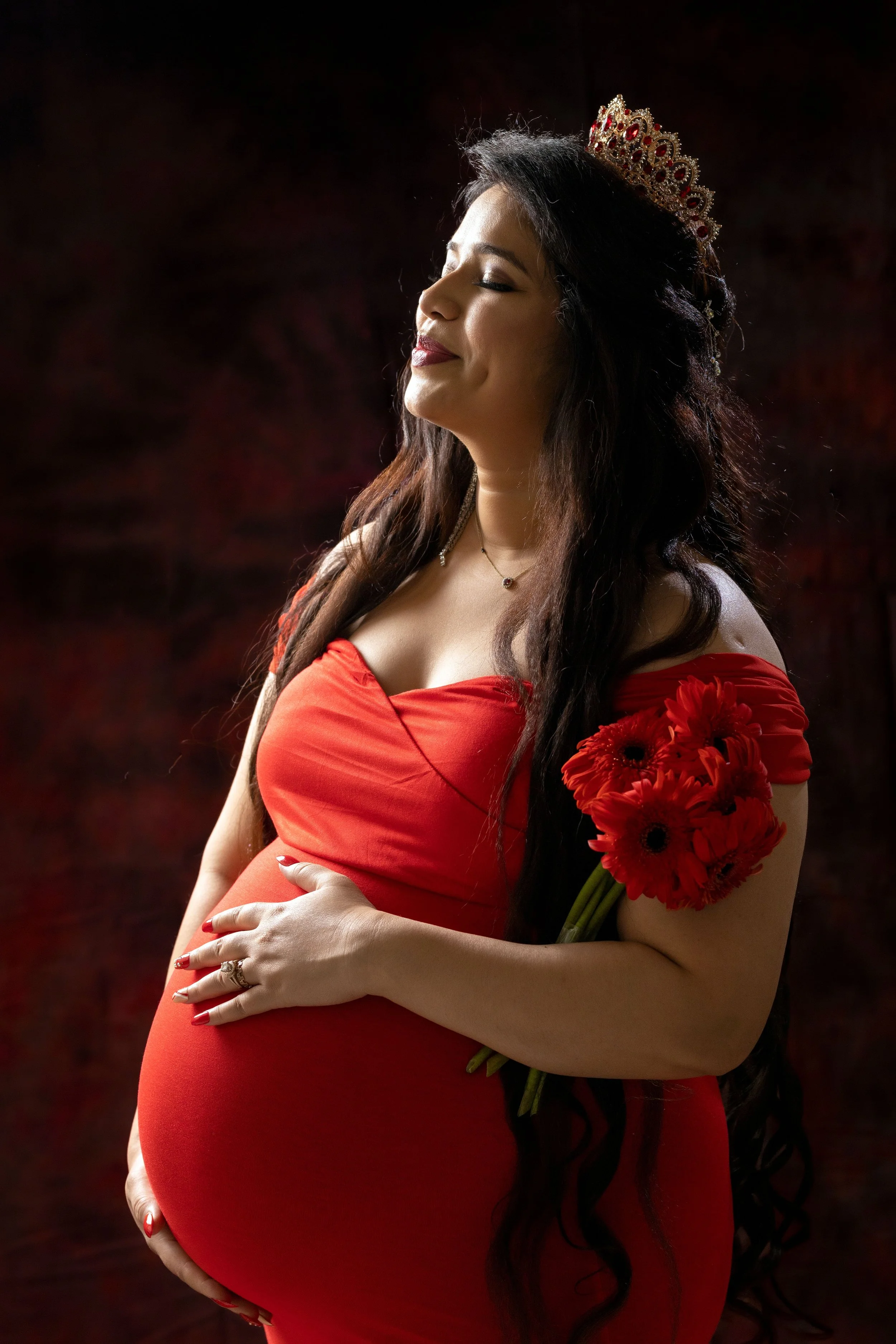A pregnant woman with long dark hair, wearing a red off-the-shoulder dress, a tiara, holding a bouquet of red flowers, and standing peacefully with her eyes closed.