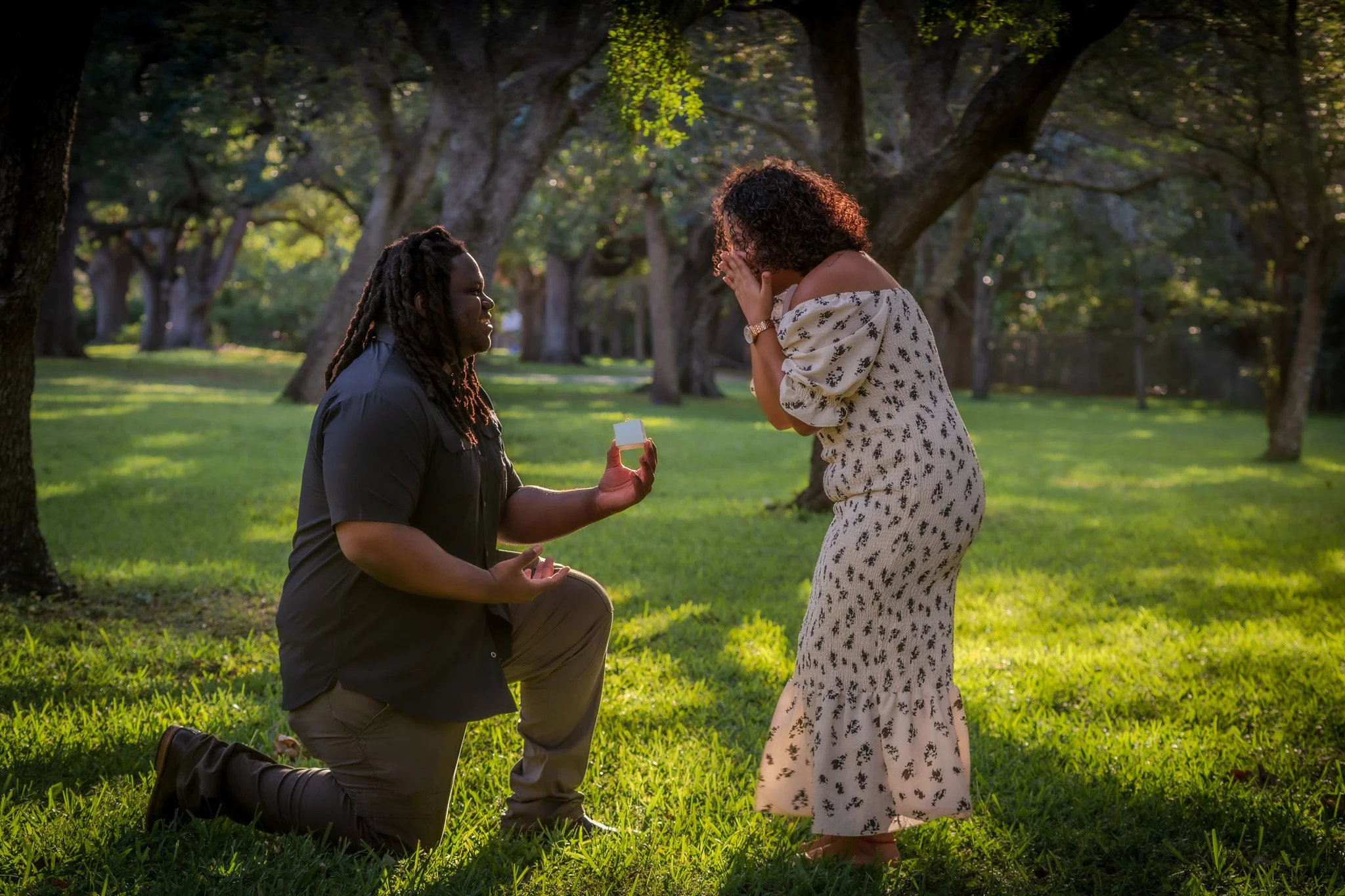 A man kneeling on one knee on a grassy field, proposing to a woman who is visibly emotional, covering her mouth with her hands. The scene takes place outdoors under a canopy of large trees in the late afternoon or early evening.