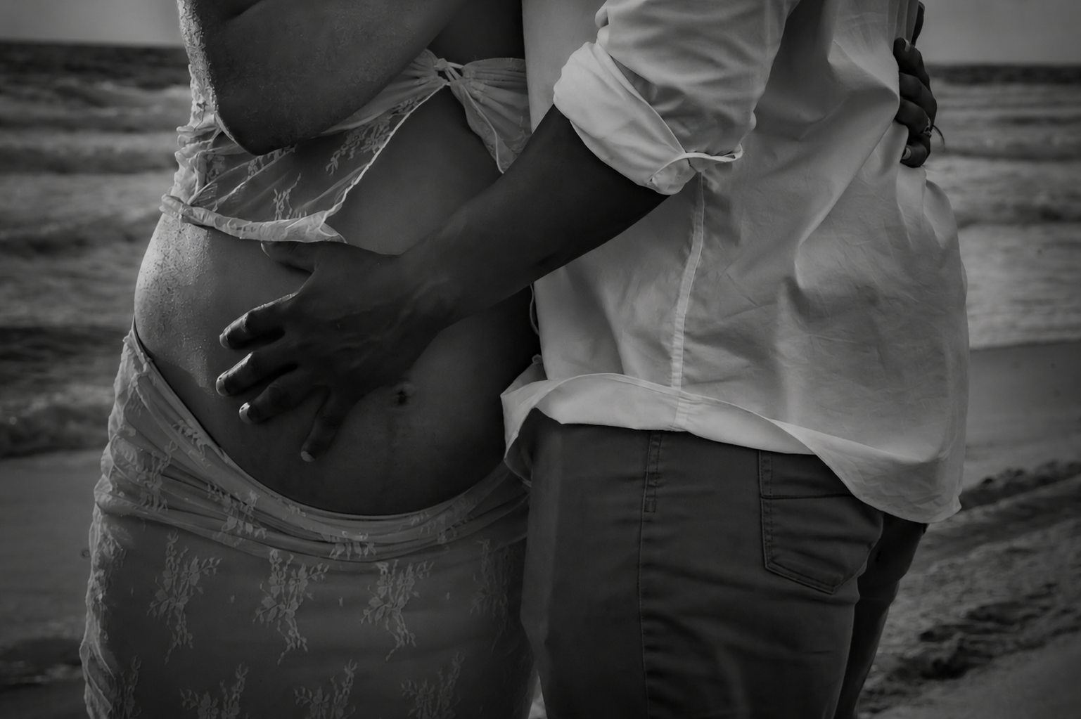 A black and white photo of a couple embracing on a beach, with the man gently holding the woman's pregnant belly.