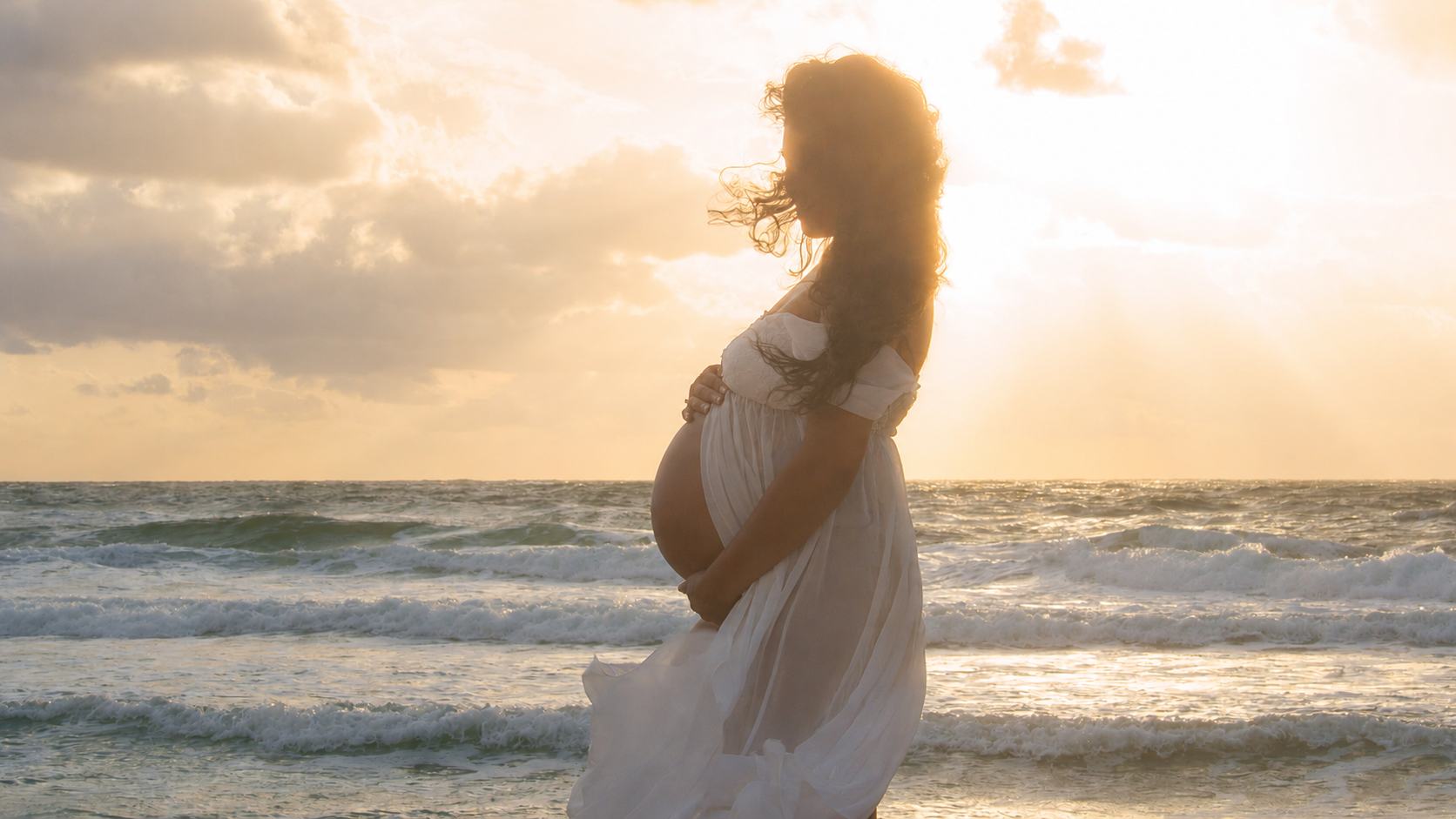 A pregnant woman in a white dress standing on the beach at sunset, with her hand on her belly and the wind blowing her hair.