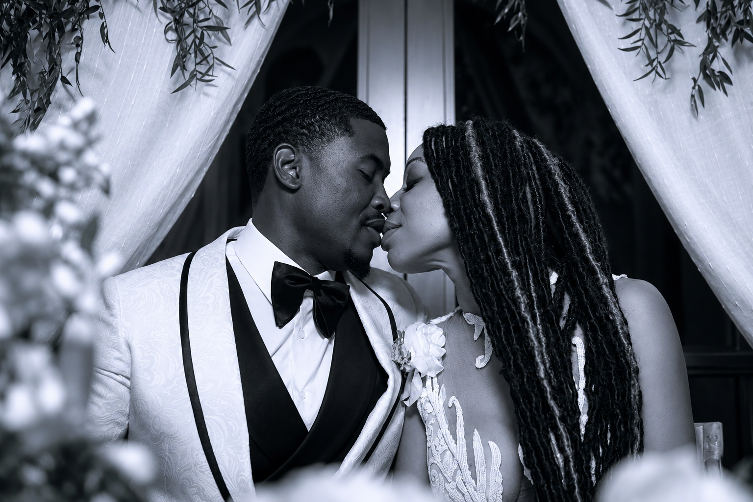 A black and white photo of a couple in wedding attire, about to kiss, with decorative curtains and greenery in the background.