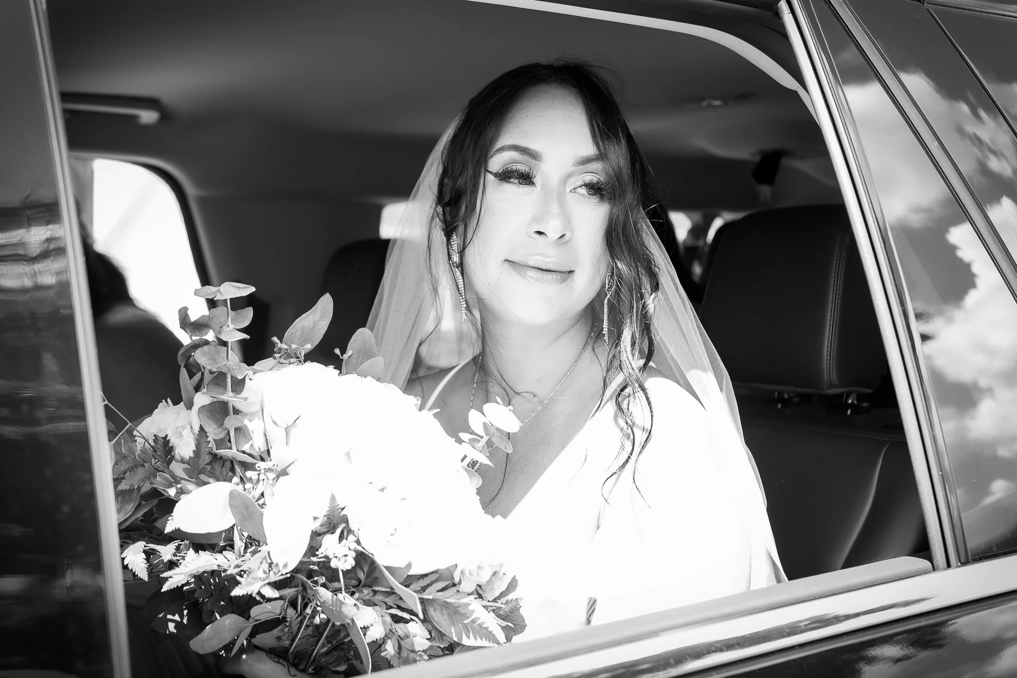 A bride with dark hair and makeup, wearing earrings and a veil, sitting in a car with a bouquet of flowers nearby, smiling softly.