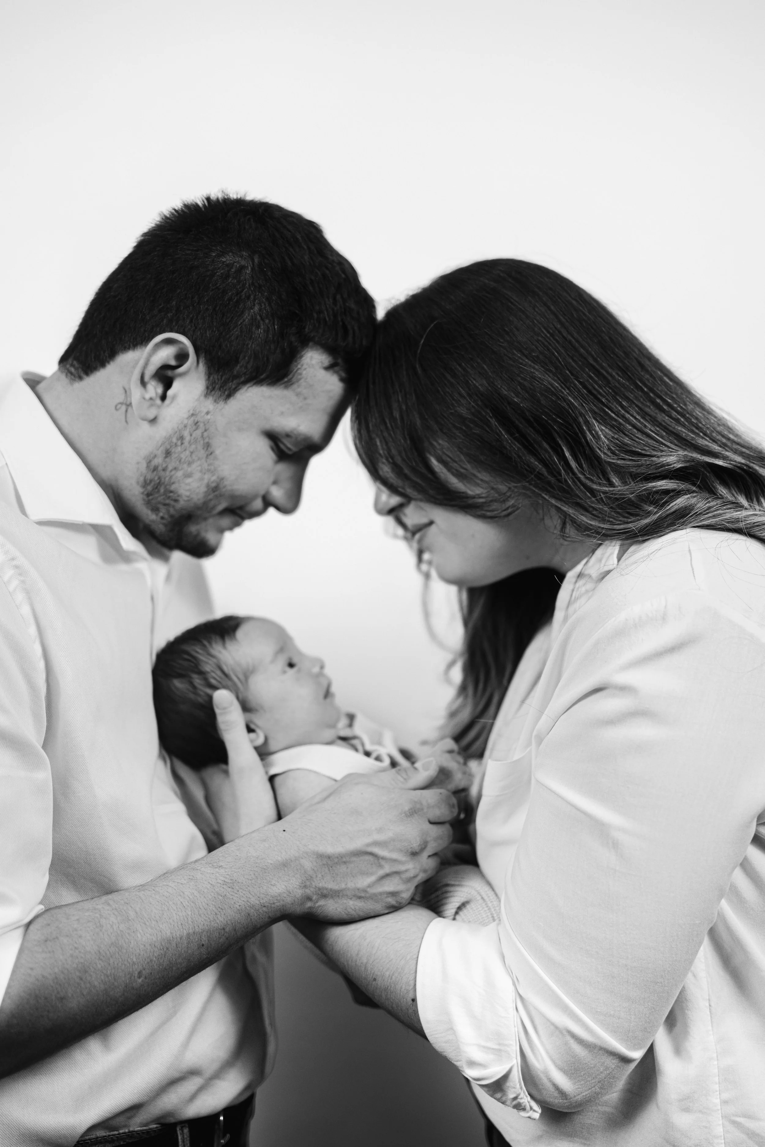 Two adults, a man and a woman, gently hold and look affectionately at a newborn baby, with their foreheads touching. The scene is in black and white.