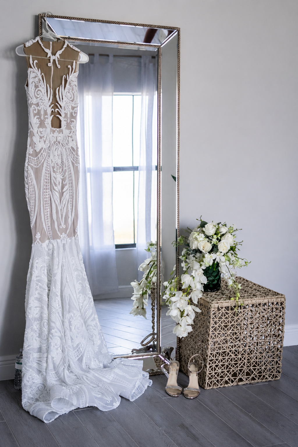A wedding dress hanging on a white hanger in front of a mirror, with white flowers and white high-heeled shoes on a woven table nearby, and a window with sheer white curtains in the background.