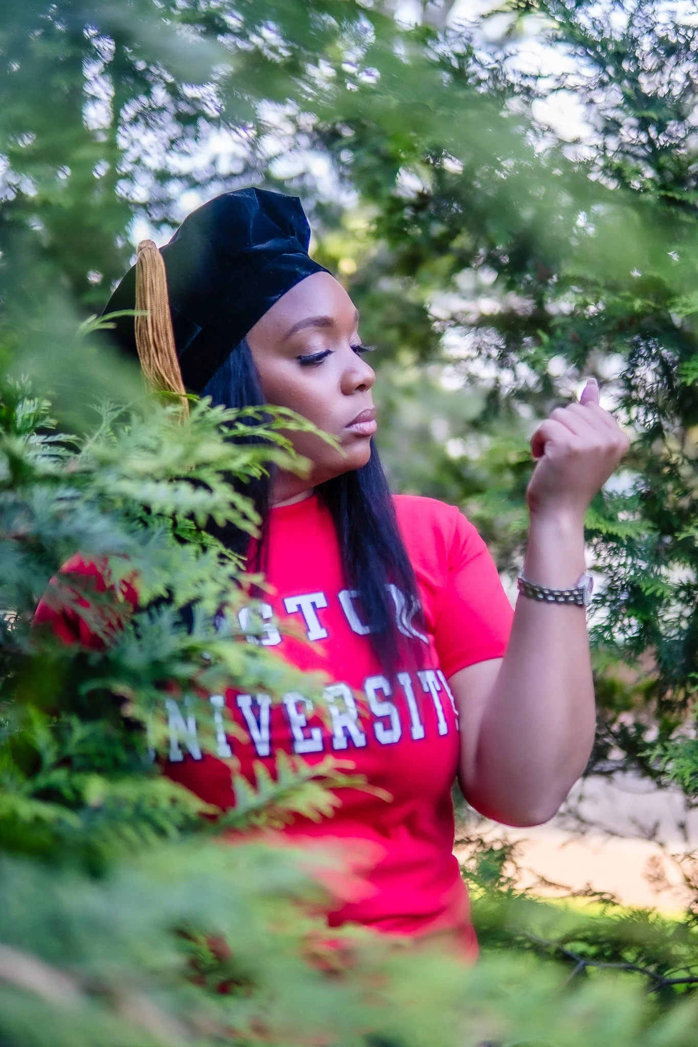 A woman with long black hair wearing a black graduation cap with a gold tassel and a red T-shirt with white and purple lettering, standing among green trees, with her eyes closed and her right fist raised.