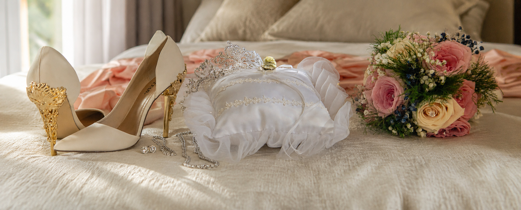 A collection of wedding accessories on a bed, including ivory high heels with gold heels, a tiara, a white pillow with gold accents, a bouquet of pink and cream roses, and a wedding ring.
