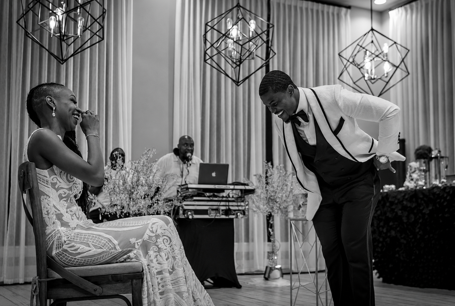 A joyful wedding reception scene with a woman in an intricate wedding dress sitting on a chair, laughing and covering her mouth with her hand, as a man in a tuxedo with a white jacket and black bow tie bows deeply to her, both smiling and sharing a h