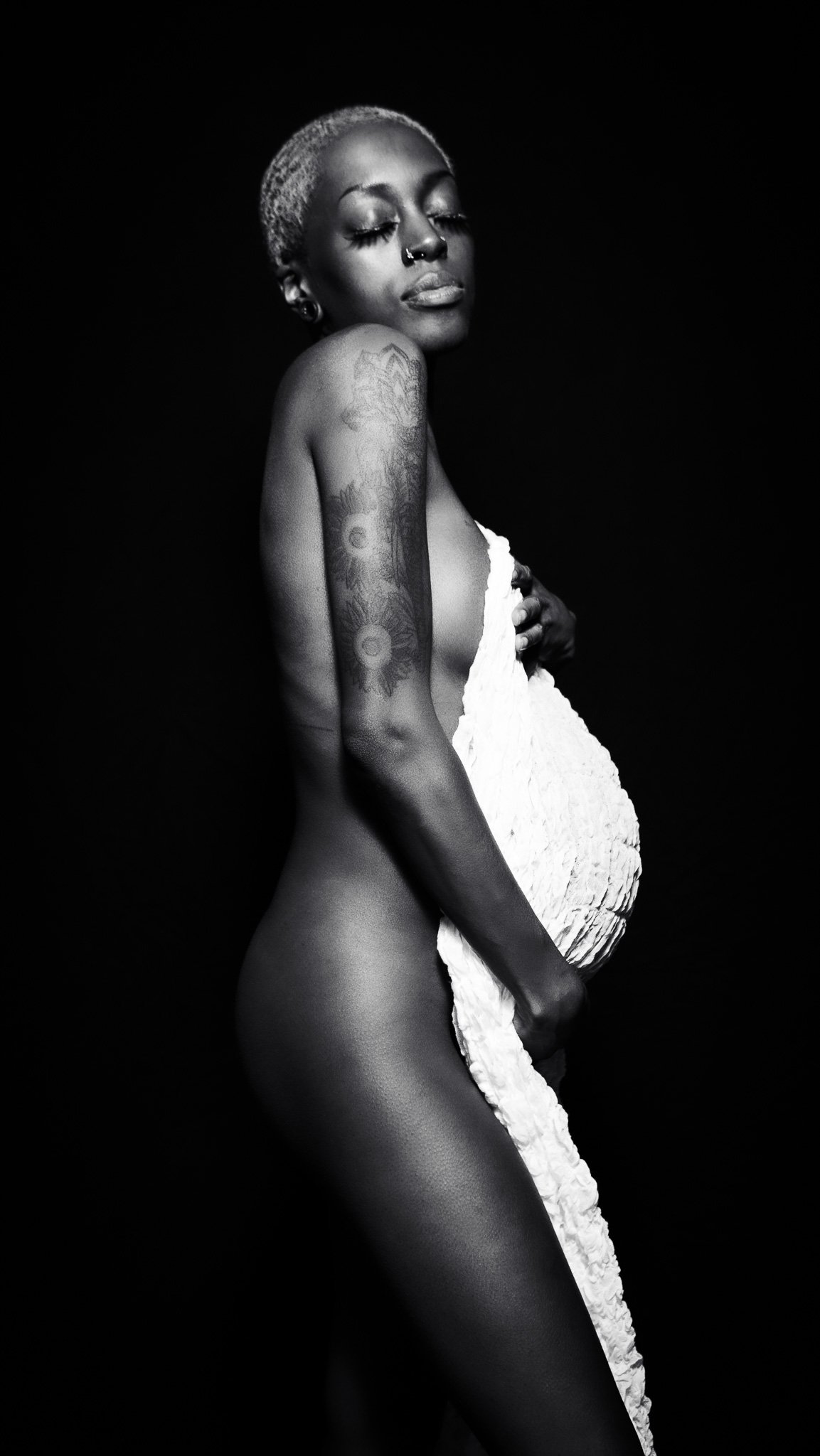 Black and white photograph of a woman with closed eyes, short blonde hair, tattoos on her left arm, and a nose piercing. She is holding a white textured cloth against her chest and side, with a dark background.