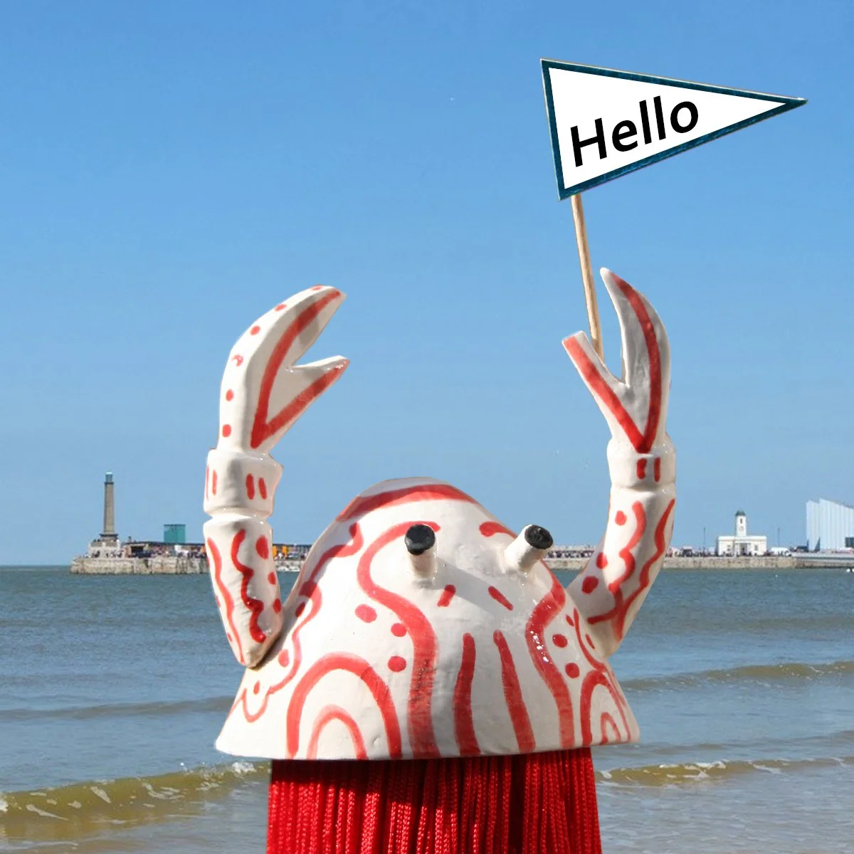 A decorative crab sculpture holding a sign that says 'Hello' with a background of a shoreline, water, and distant buildings.