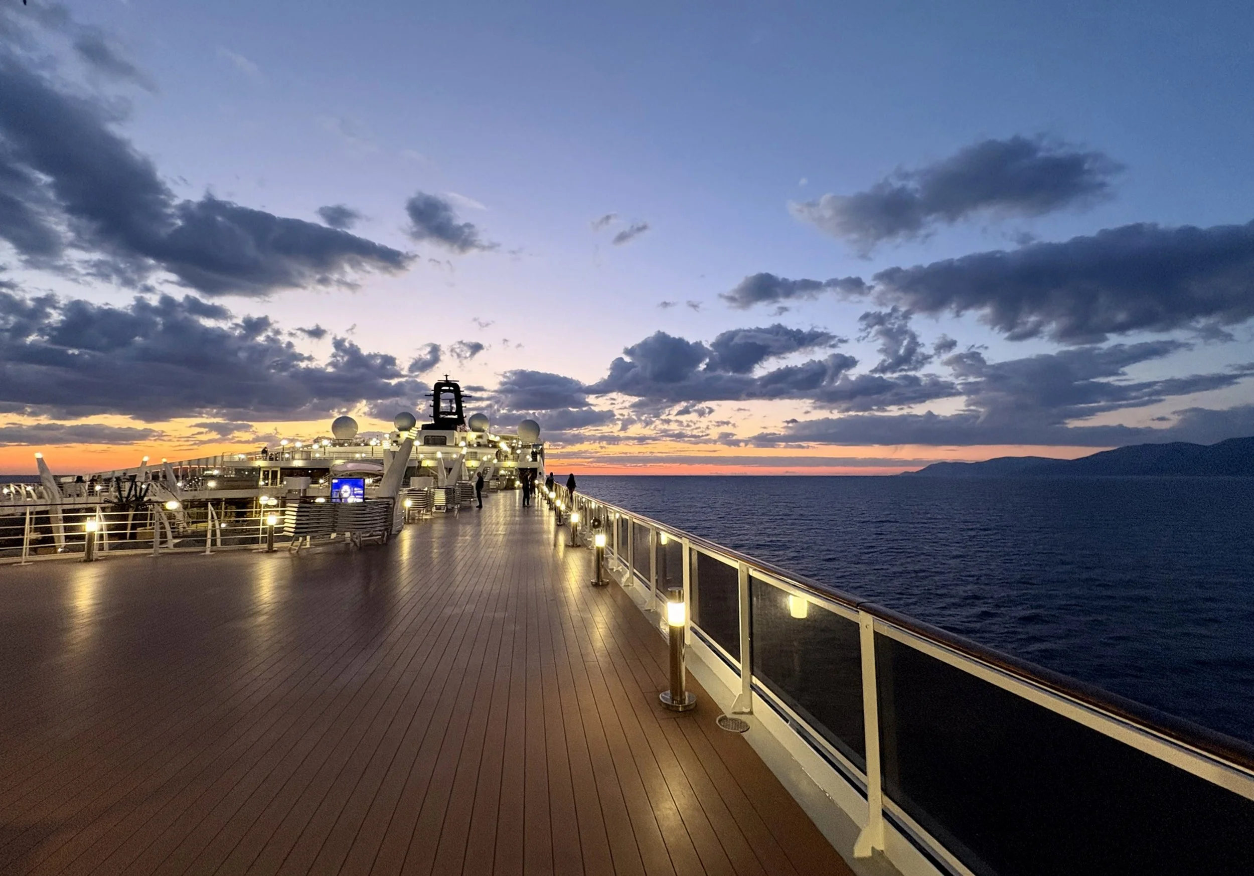 View of the deck of a cruise ship at sunset with clouds in the sky and the ocean in the background.