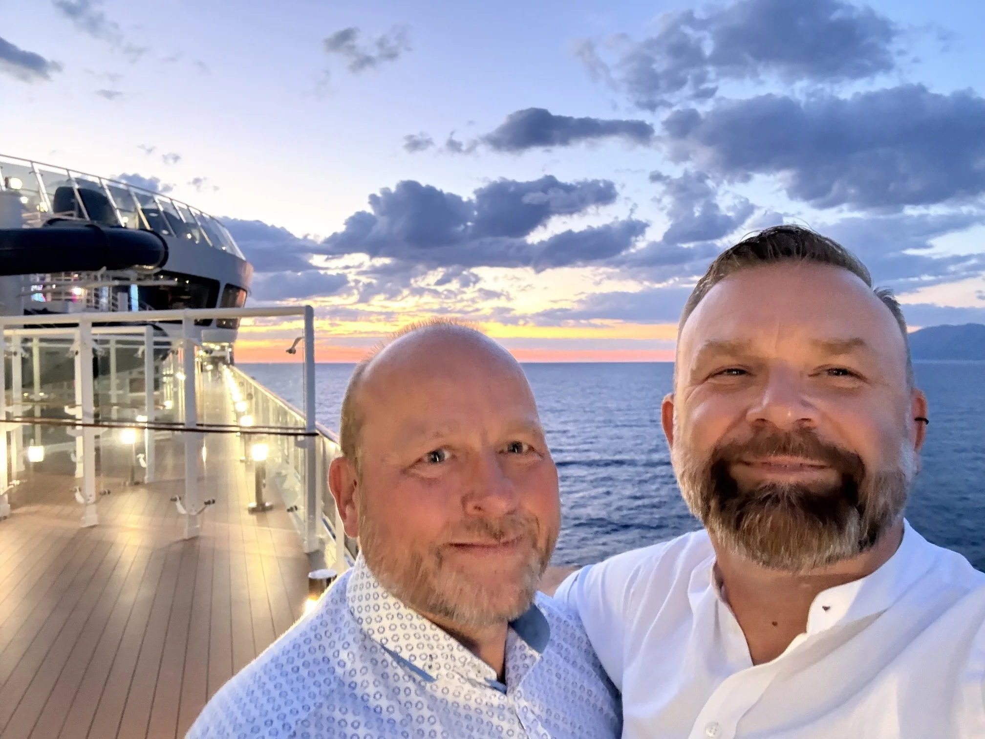 Two men smiling taking a selfie on a ship deck during sunset with partly cloudy sky over the ocean.