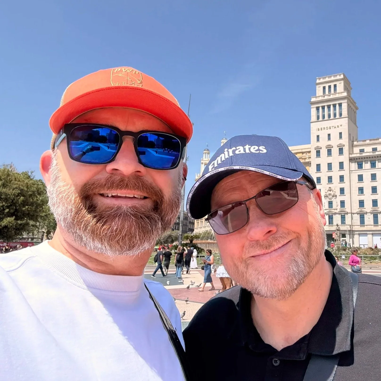 Two men taking a selfie in a city park with a historic building in the background. One man has a beard and is wearing sunglasses, a white shirt, and a red cap. The other man has a goatee, is wearing glasses, a dark shirt, and a blue cap with the word "Emirates" on it.