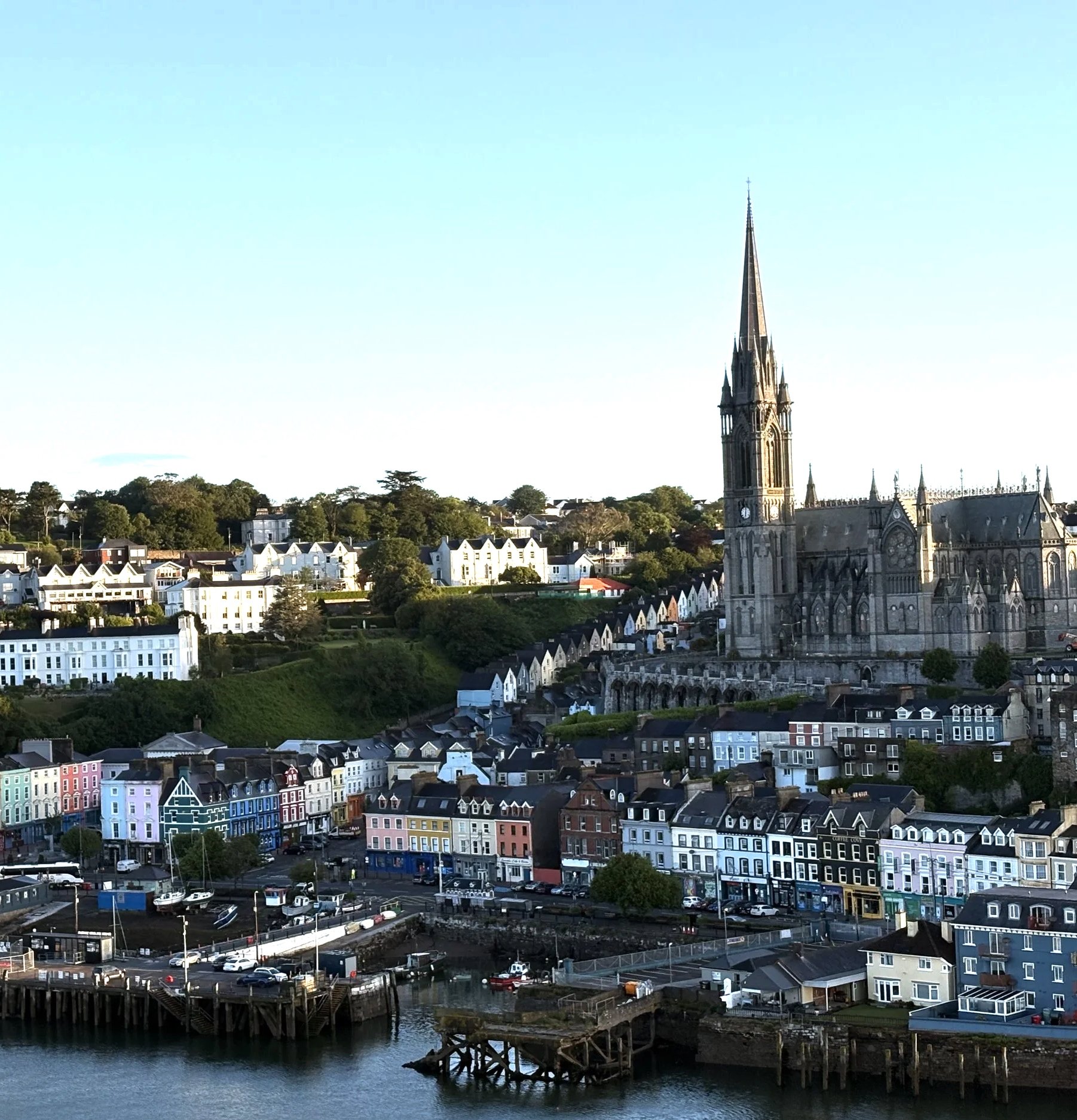 A coastal town with a large church in the background and colorful houses along the waterfront. The church features a tall spire, and there are boats docked by the pier, with water and trees surrounding the area.