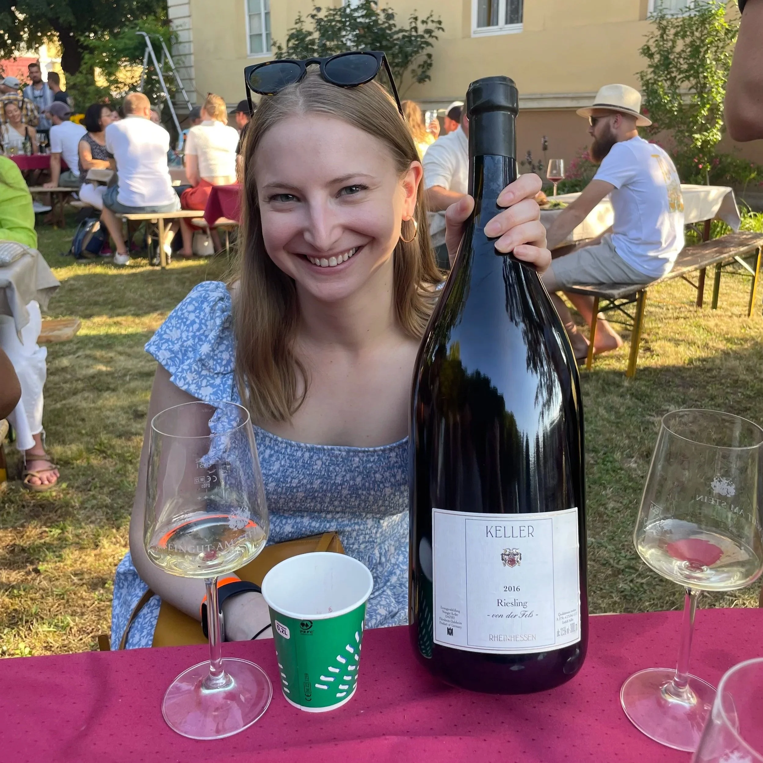 A close-up photo of a smiling woman holding a dark wine bottle at an outdoor wine tasting event. The woman has long blonde hair, is wearing a blue patterned dress, and also has sunglasses resting on her head. In front of her are empty wine glasses and a green paper cup. In the background, people are seated at tables and socializing, with greenery and a yellow building visible.