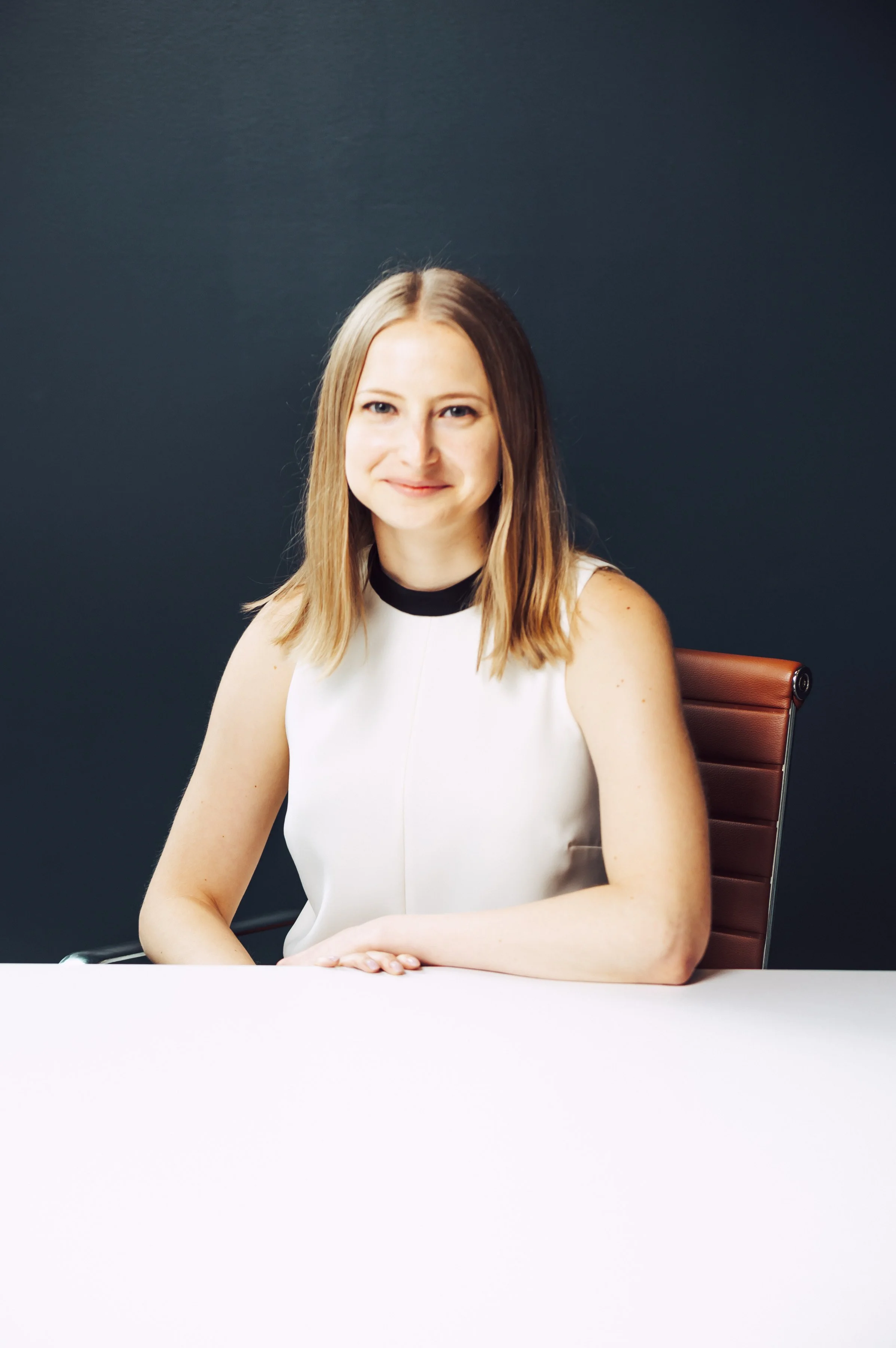 A woman with shoulder-length blonde hair, wearing a sleeveless white top with a black collar, sitting at a conference table with her hands resting on the table. She is smiling slightly and sitting in front of a dark background with a brown chair visible behind her.