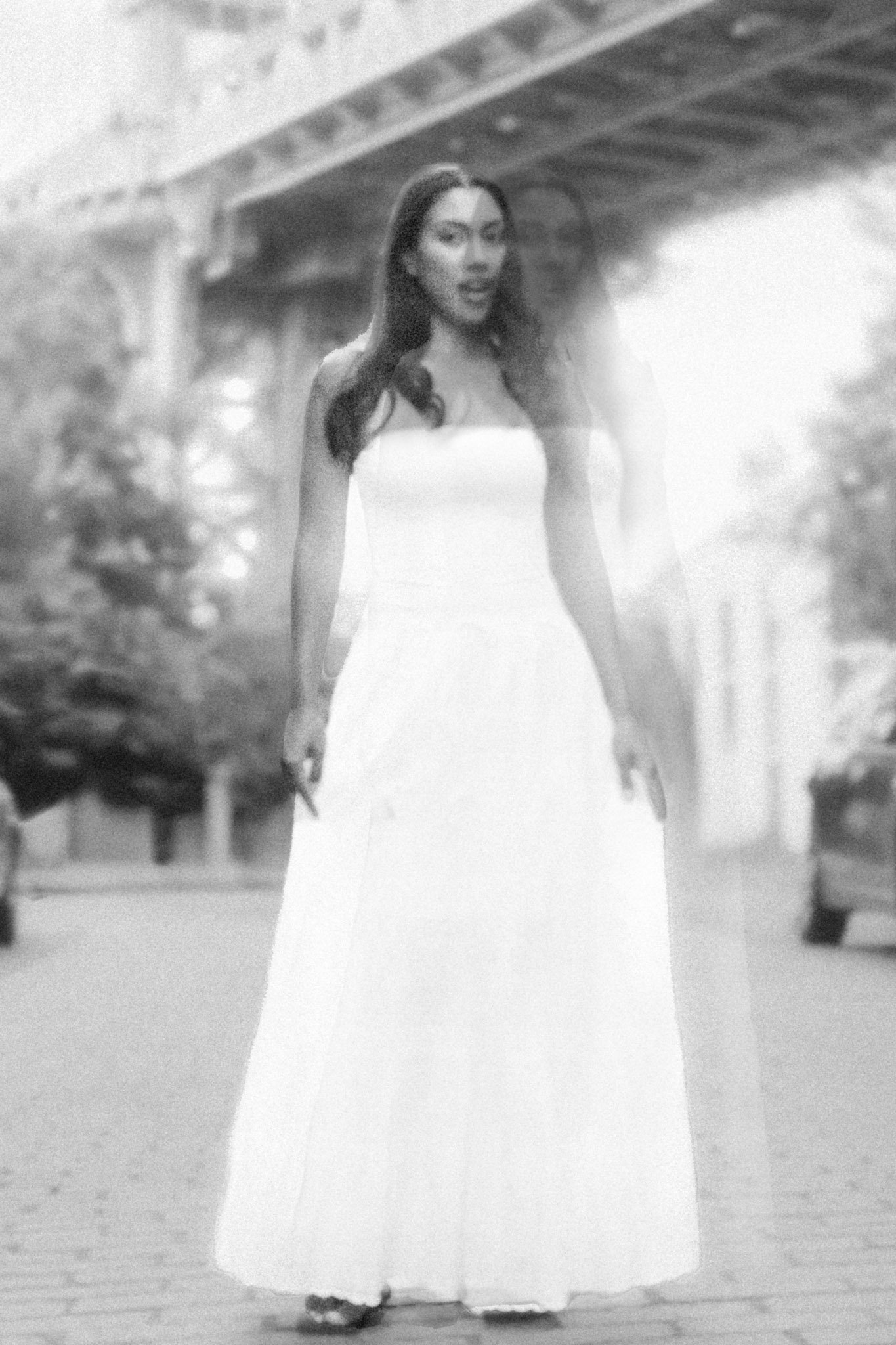 A woman in a strapless white dress standing outdoors on a brick-paved street with cars and trees in the background.
