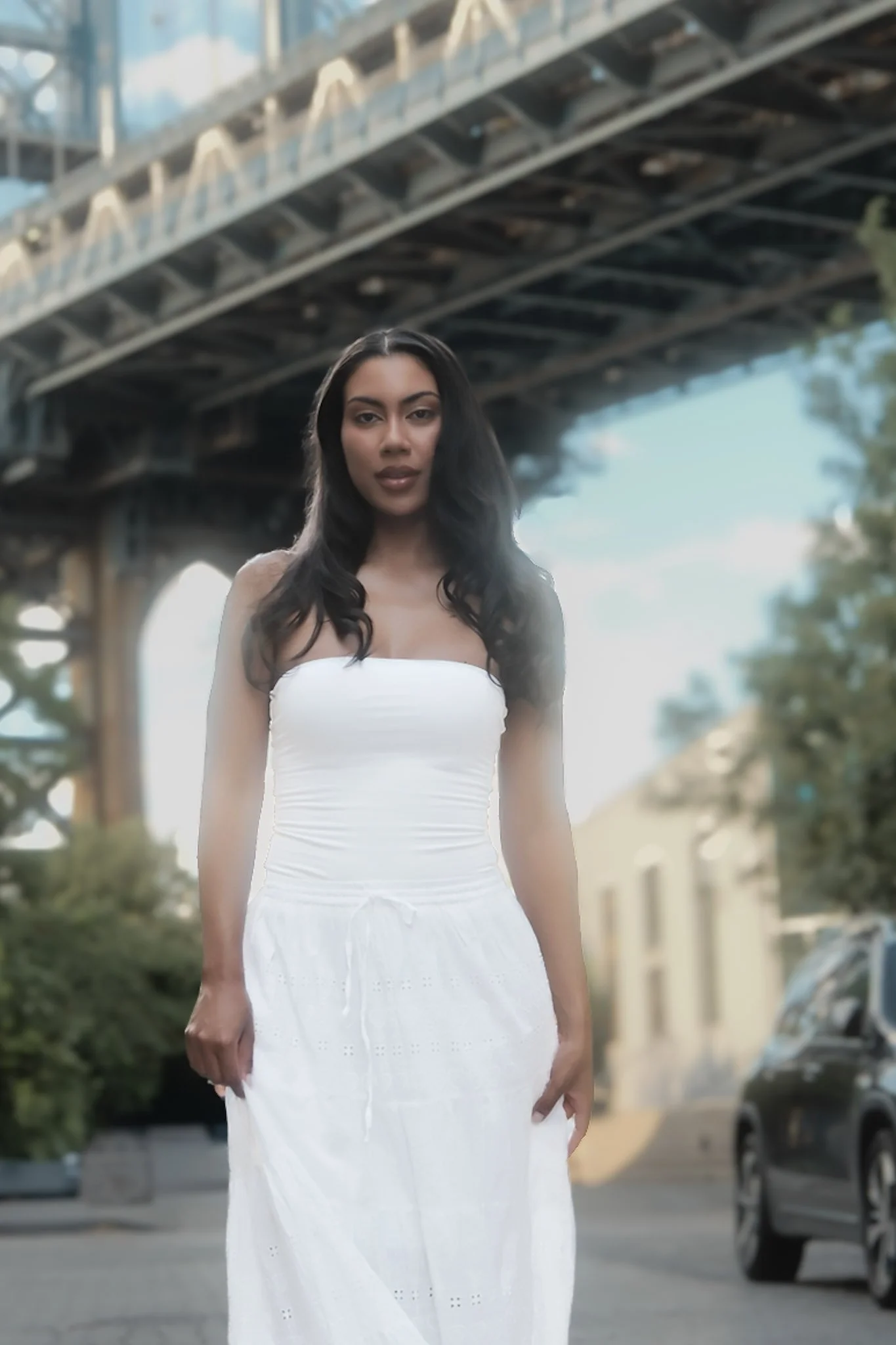 A woman with long dark hair standing outdoors under a bridge, wearing a strapless white dress.